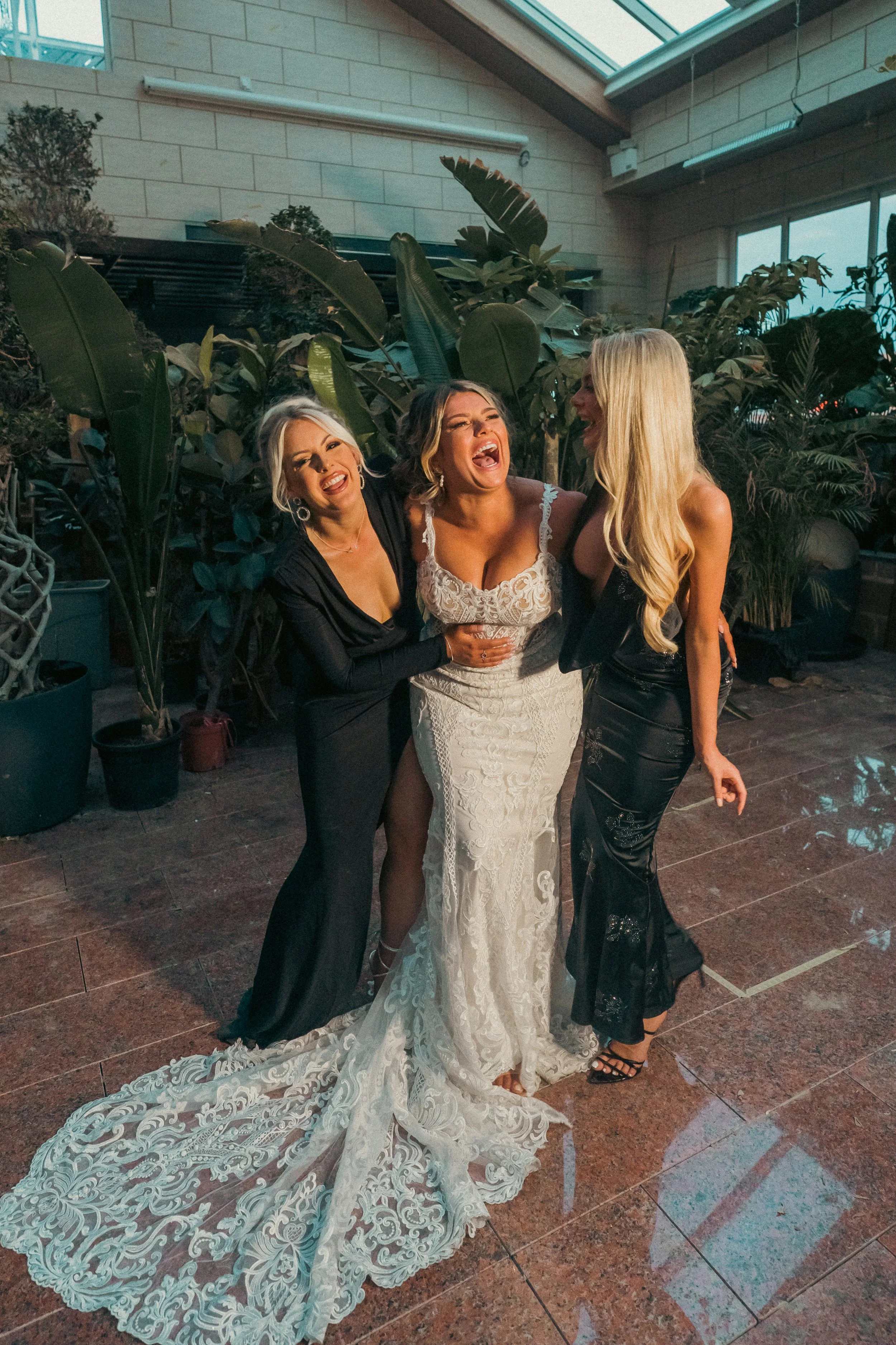 Three women in elegant evening wear laughing and enjoying themselves indoors, with large green plants and a glass ceiling above.