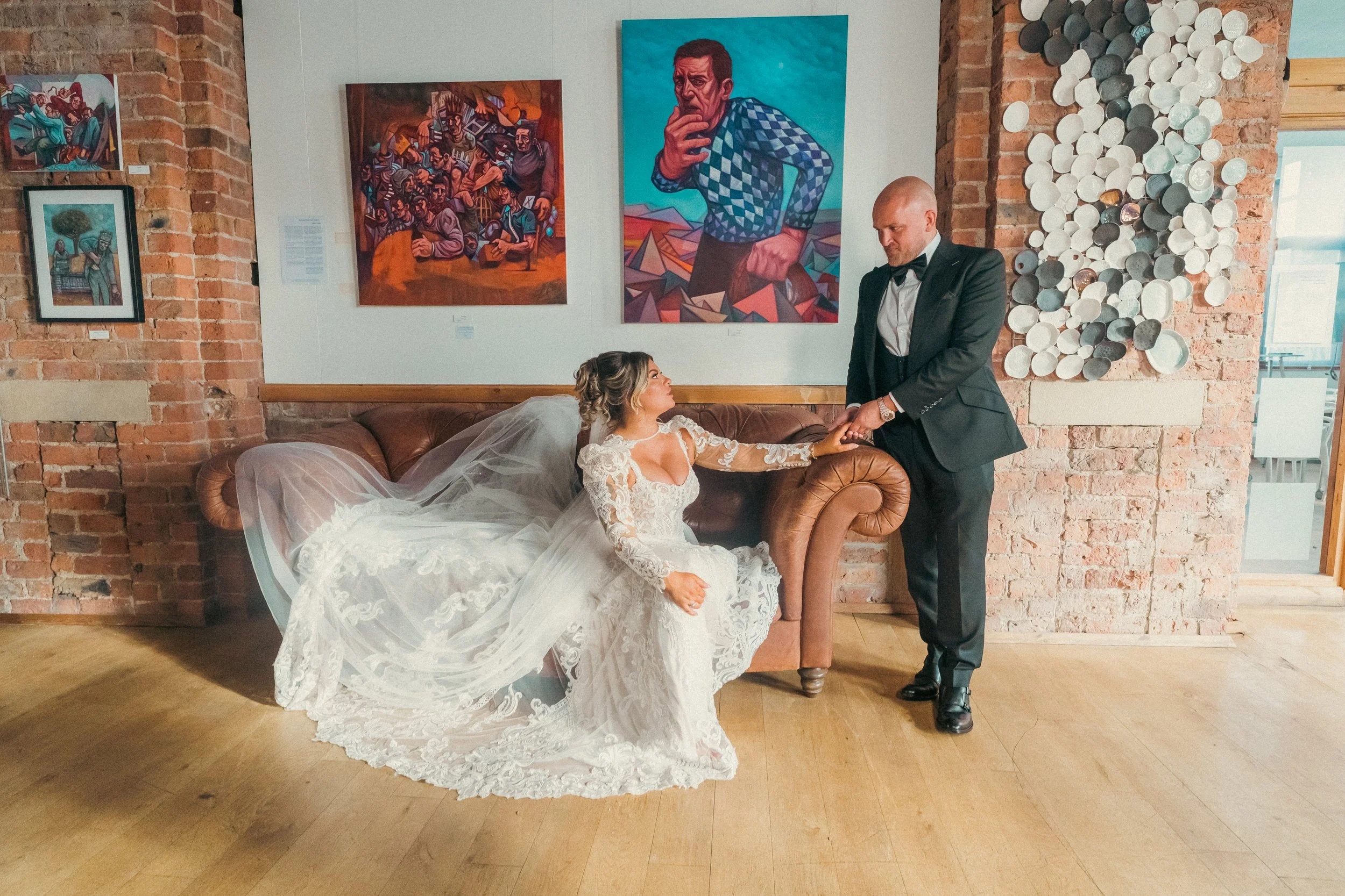 A bride in a lace wedding dress and veil is sitting on a brown leather couch, reaching out to hold hands with a groom in a tuxedo who is standing next to her. The setting appears to be an art gallery with brick walls decorated with colorful paintings