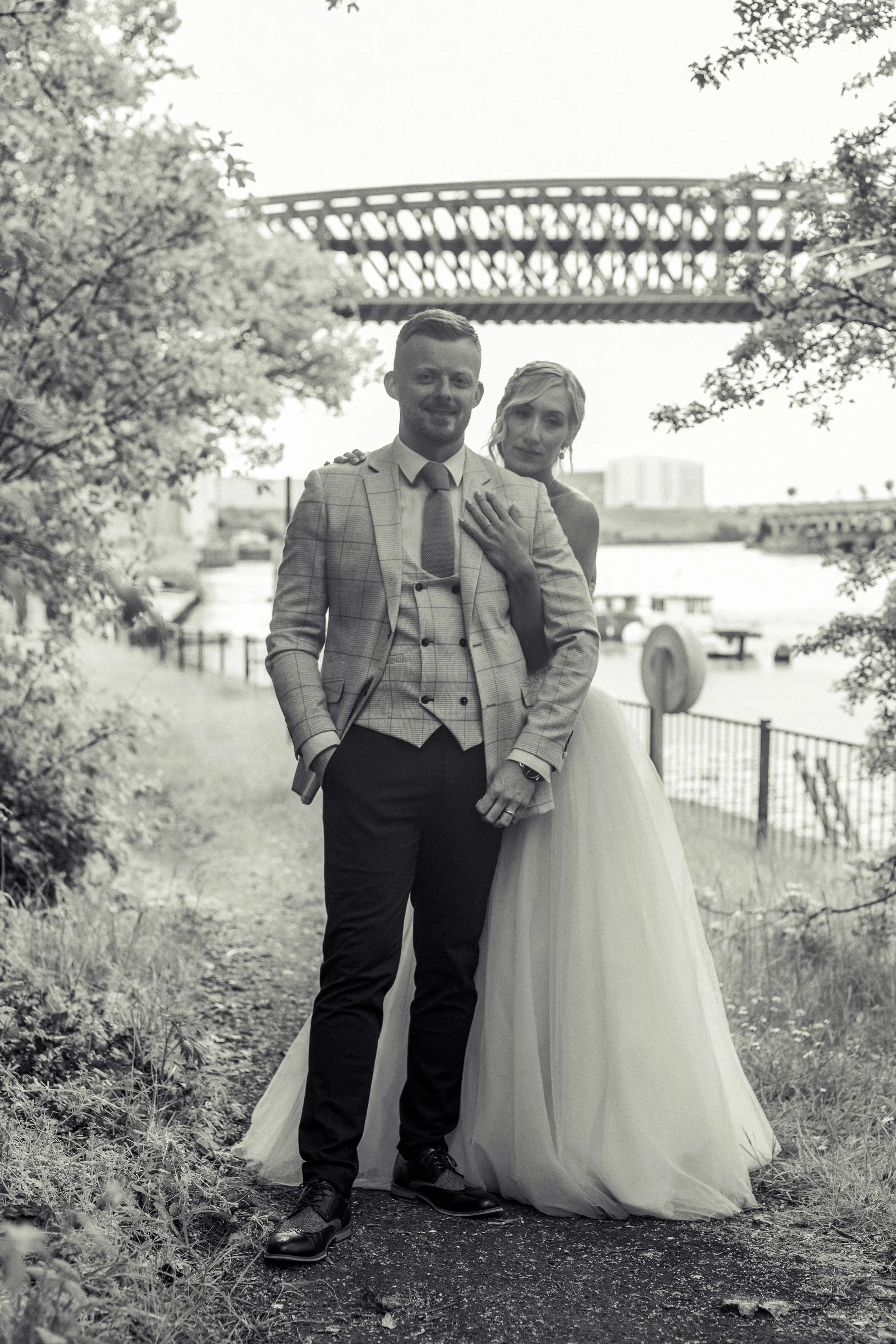 A black and white photo of a couple standing outdoors near a waterfront; the woman is in a wedding dress with her hand on the man's shoulder, and the man is in a suit, both smiling.