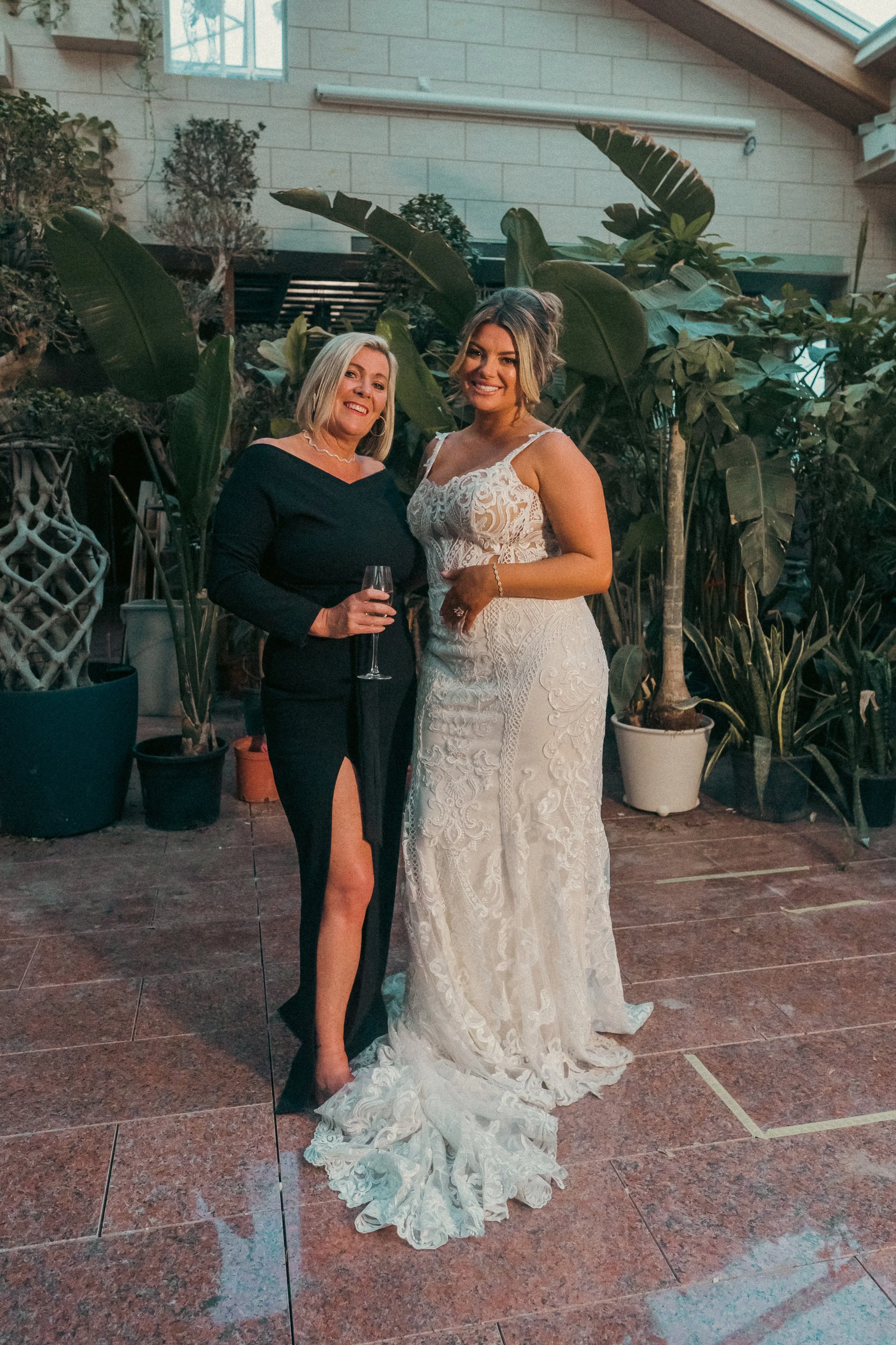 Two women standing together, one in a black dress with a slit and one in a white lace wedding dress, posing in front of potted plants indoors, smiling.
