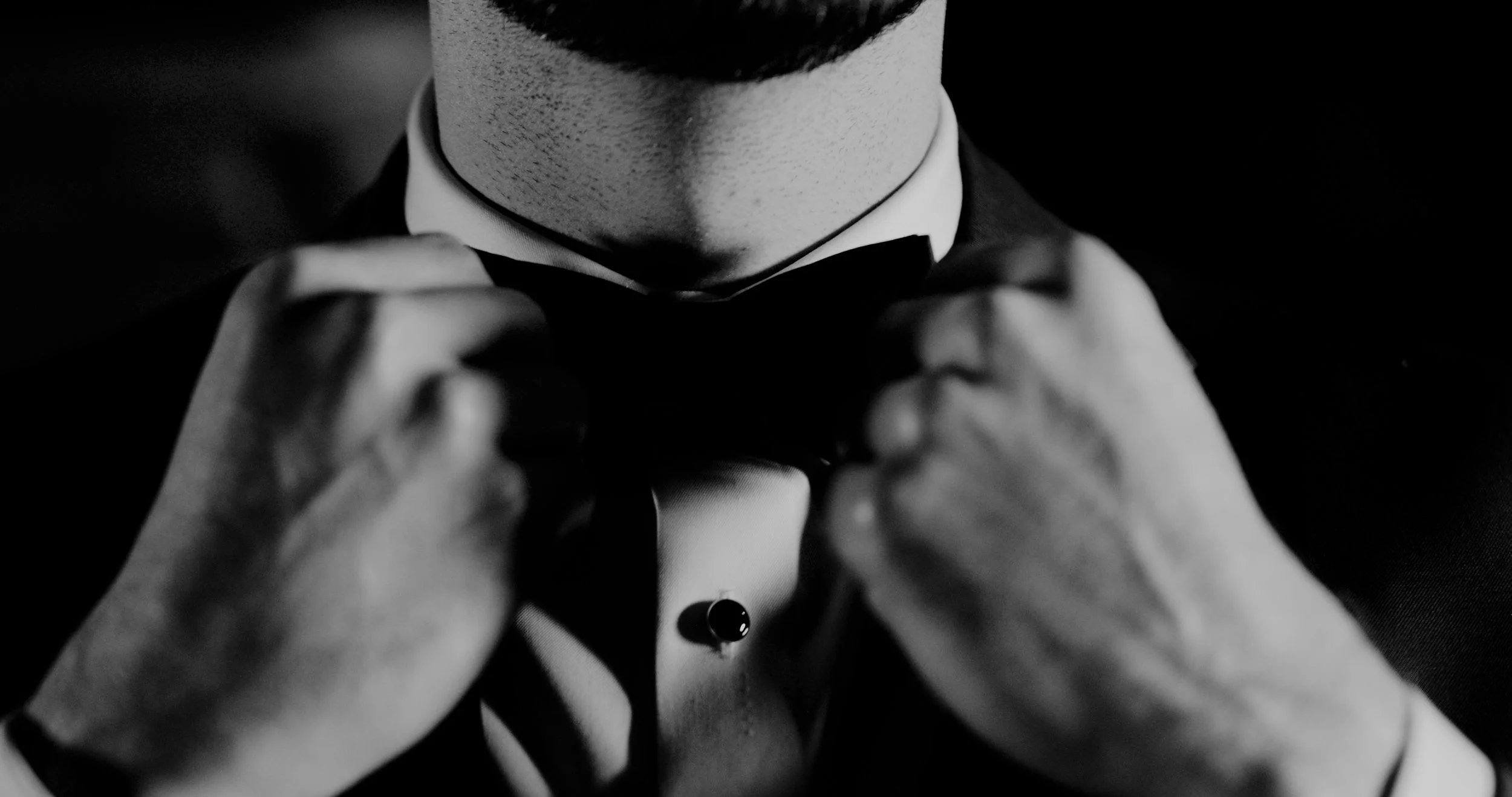 A person adjusting a black bow tie, dressed formally in a tuxedo, with focus on the hands and lower face, black and white photo.