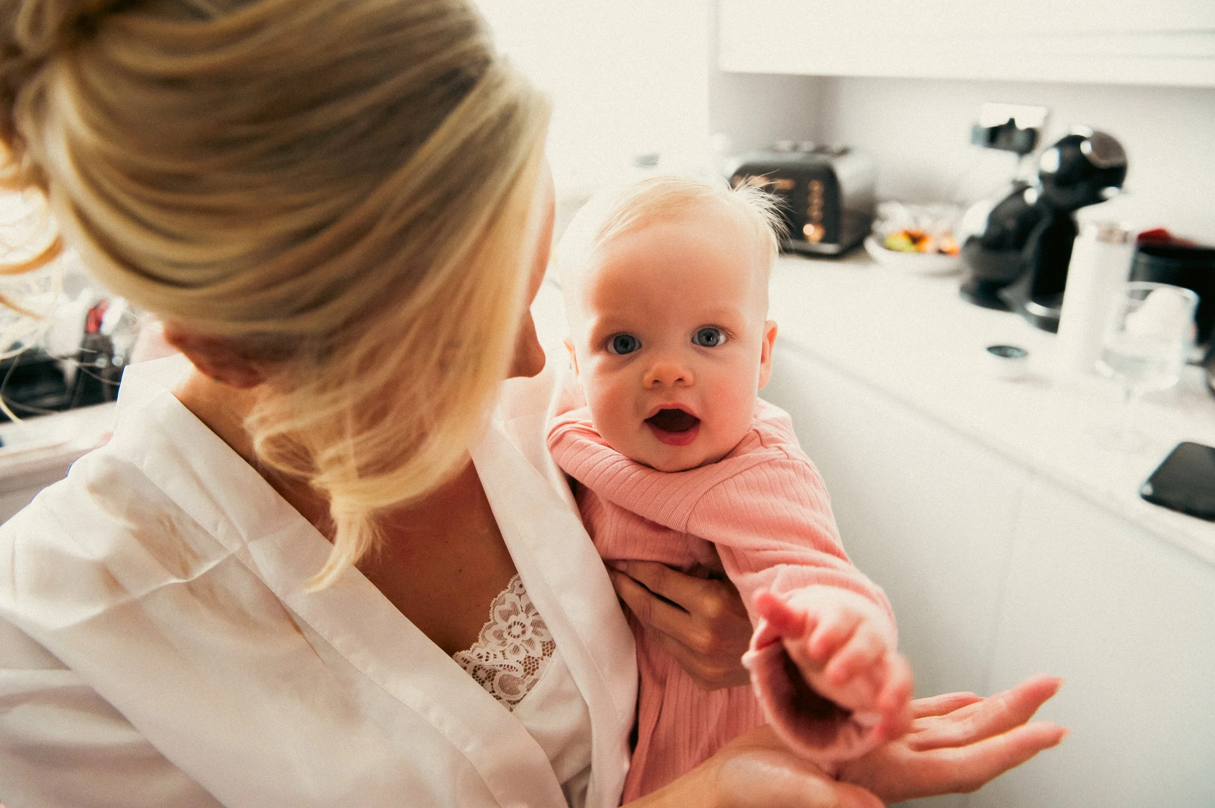 A woman with blonde hair holding a baby girl with blue eyes and light blonde hair in a pink outfit, inside a kitchen