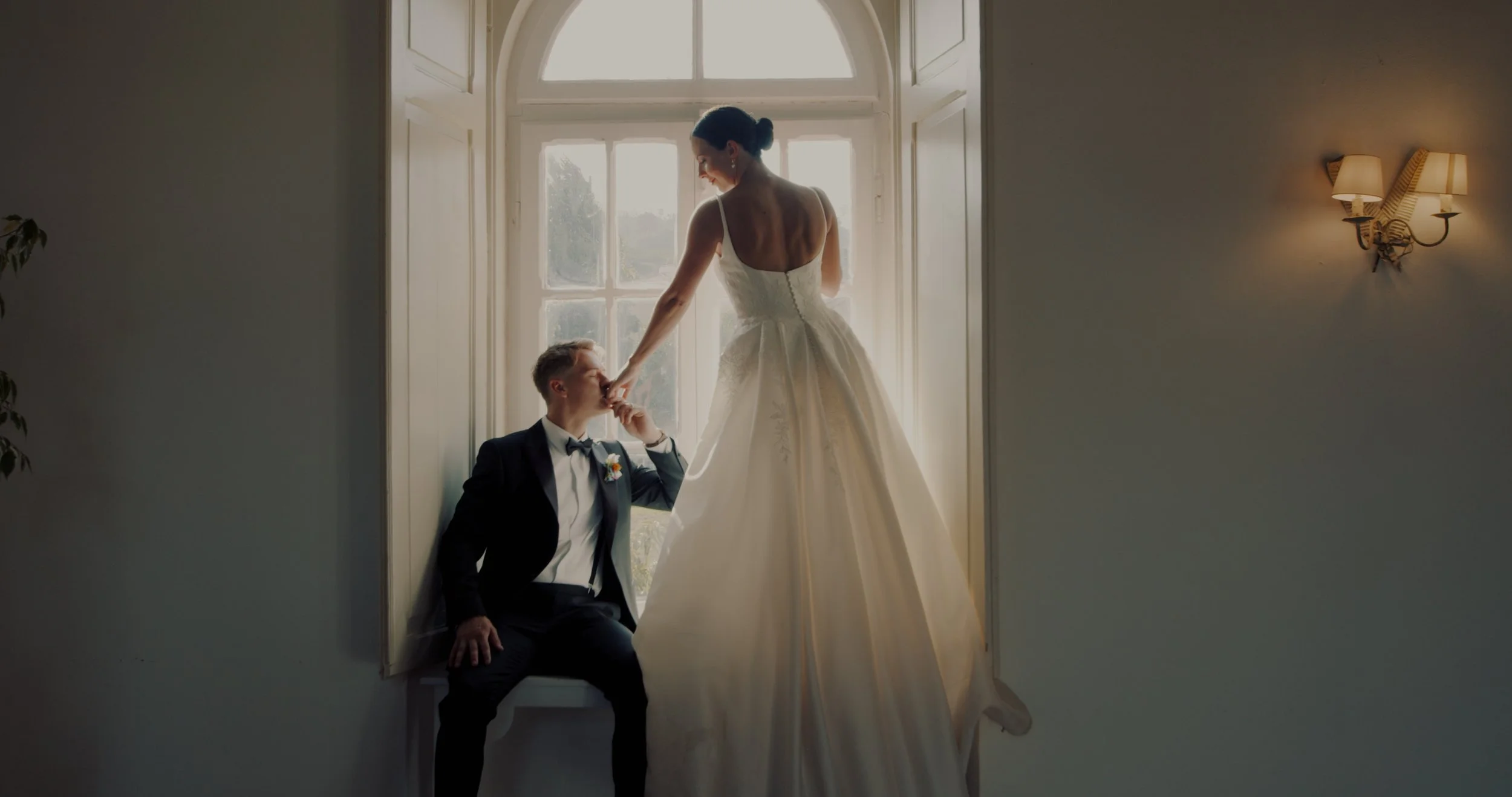 A bride standing on a window ledge, gently holding the hand of a groom seated below her, in a warmly lit room with curtains and wall sconces, during a wedding photoshoot.