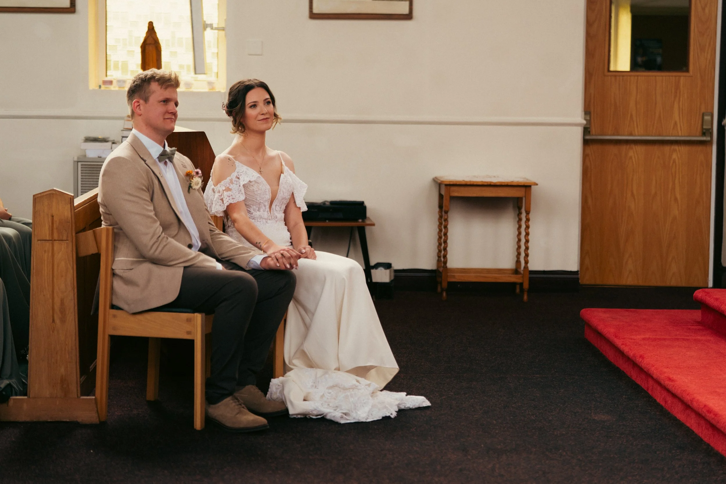 A bride and groom sitting on a wooden bench during their wedding ceremony, holding hands, in a church with a beige interior and red carpeted steps.