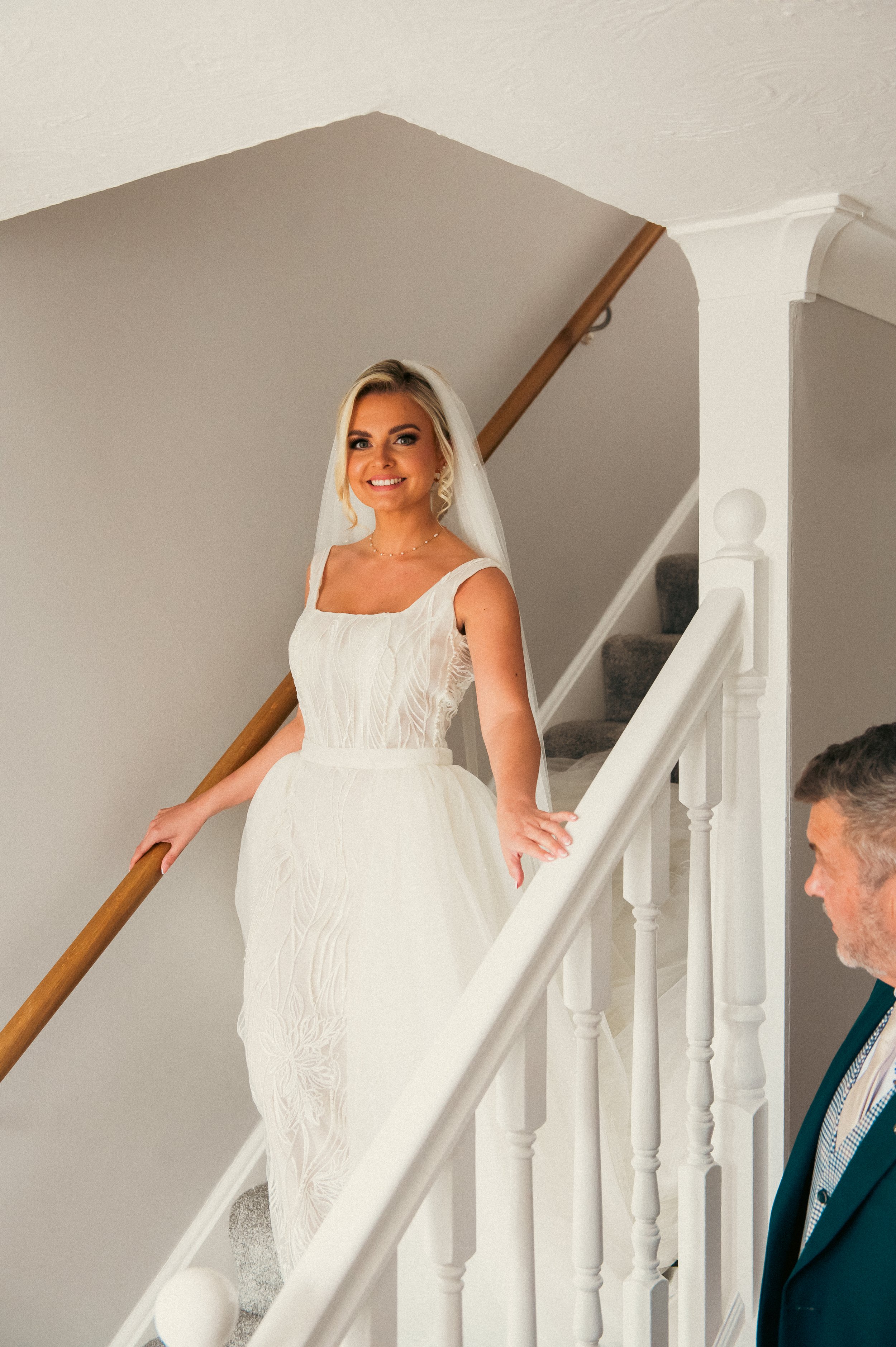 A bride in a white wedding dress and veil standing on stairs indoors with a smiling man nearby.