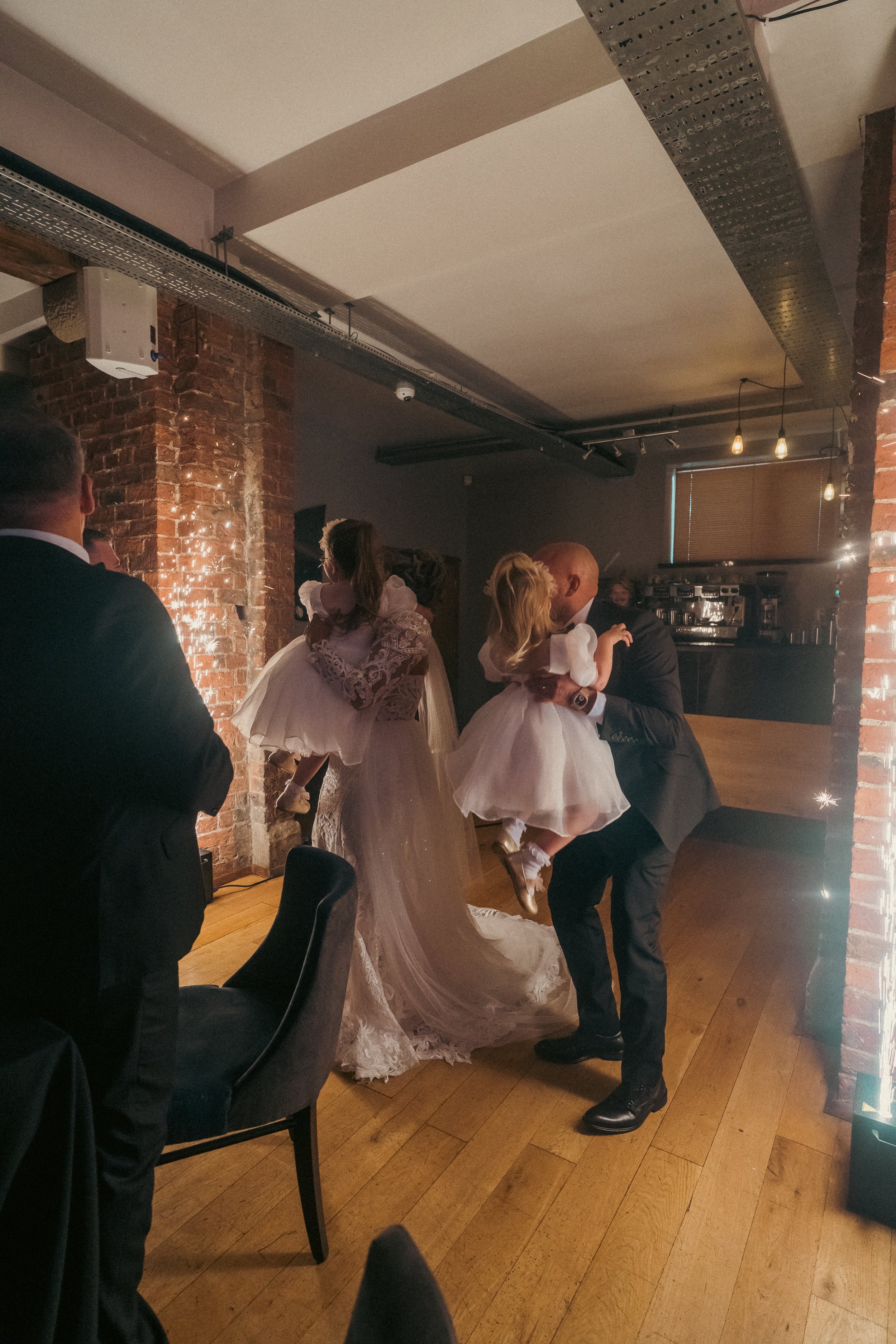 A man in a suit and a woman in a wedding dress exchanging rings during a wedding reception, with children dressed in white dresses and other guests watching.