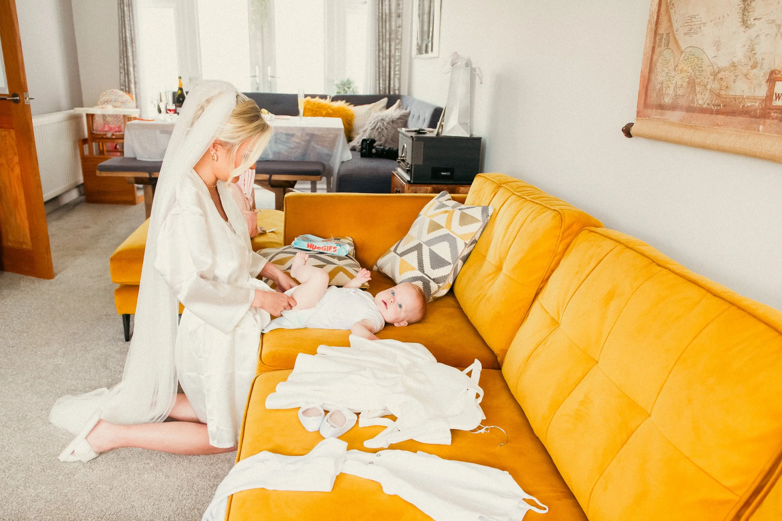 A woman dressed in a satin robe and a veil kneels on a beige carpet, changing a baby's diaper on a mustard yellow sofa. The baby, lying on its back, looks up at her. Baby diapers and wipes are on the sofa, with some baby clothes and socks nearby. The