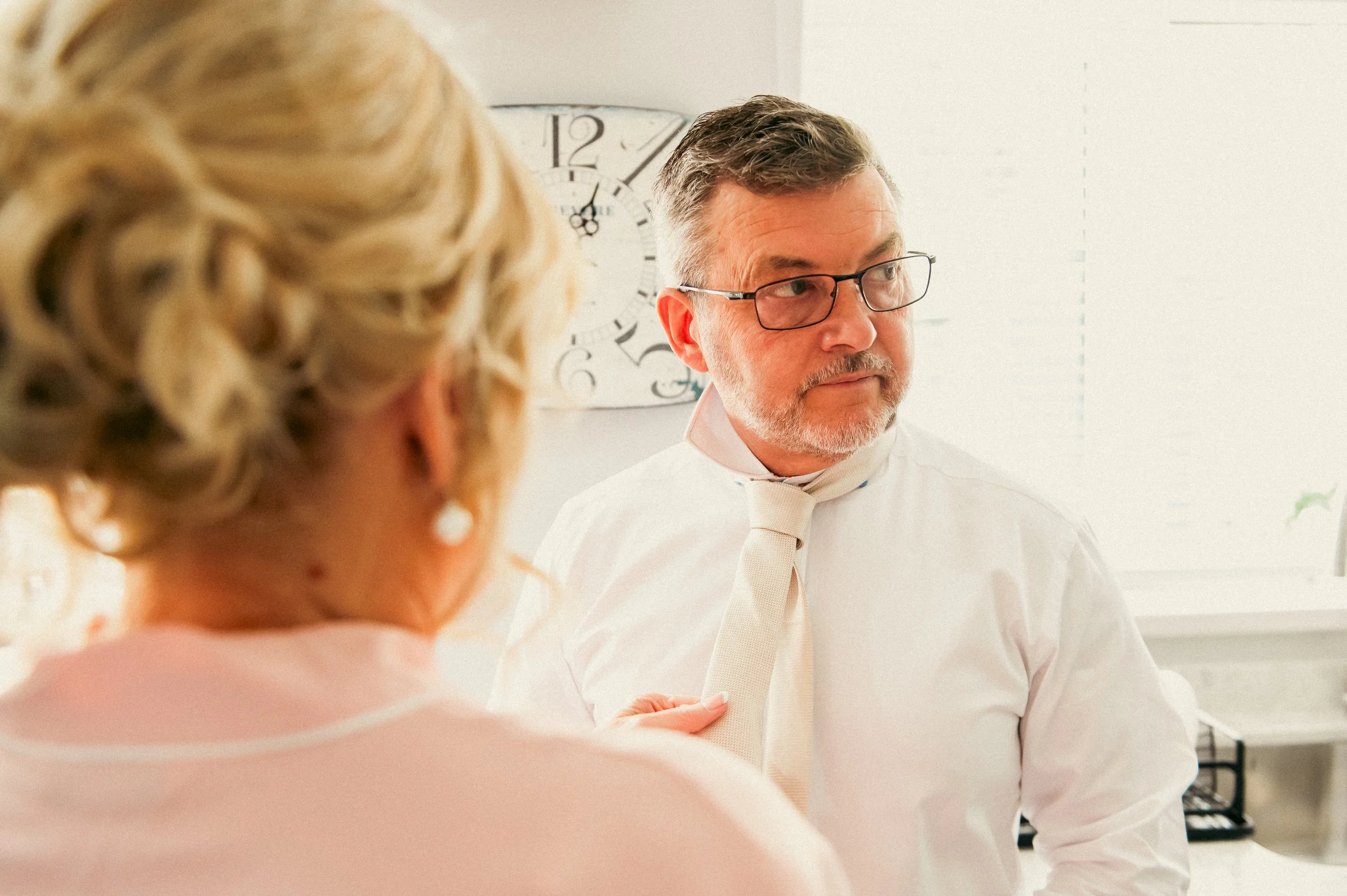 A man with glasses and a beard looks thoughtfully while a woman, whose face is not visible, adjusts his tie in an office. There is a clock on the wall behind them.