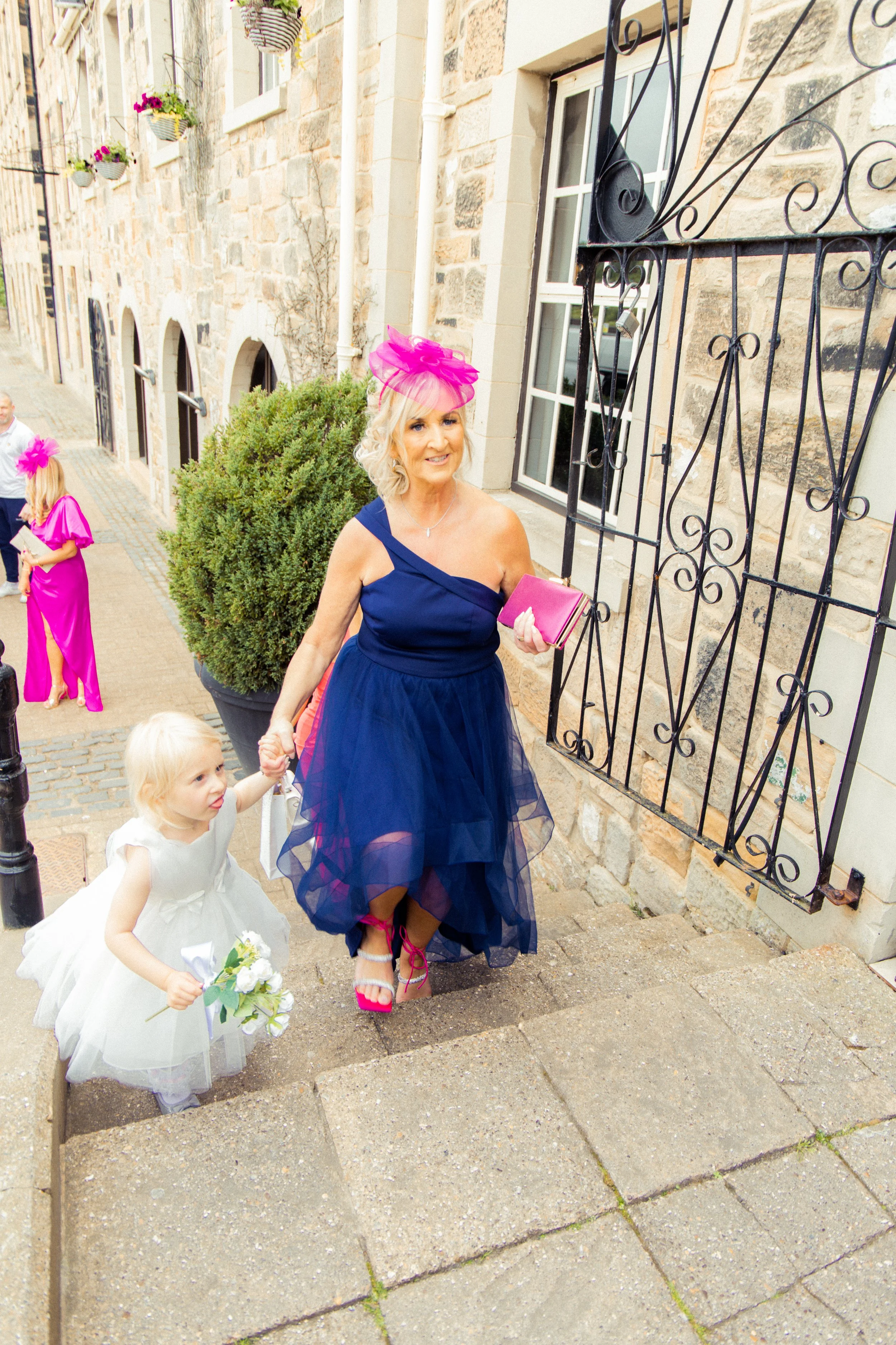 A woman in a blue dress and pink shoes, wearing a pink fascinator, walking up the steps holding a pink clutch bag and hand of a young girl in a white dress with flowers, outside a stone building with flowers on the windows.