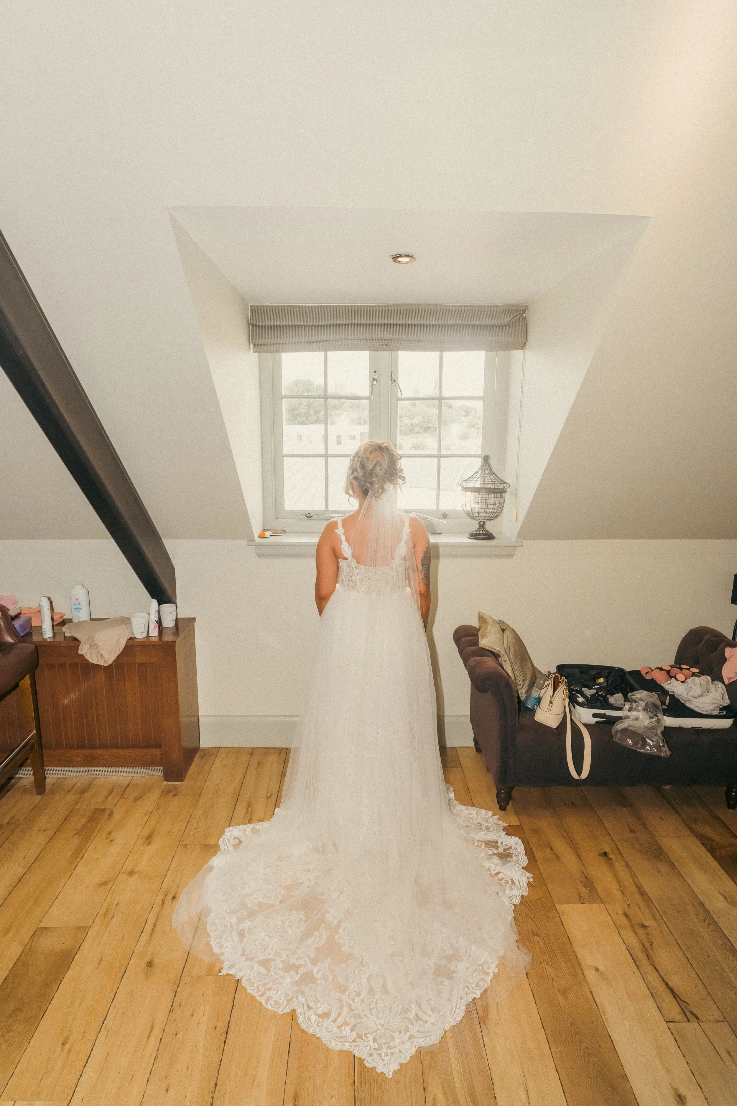 A bride in a white wedding dress standing indoors, facing a window with her back to the camera, with a wooden floor and a small bench on the left and a dark brown couch on the right.