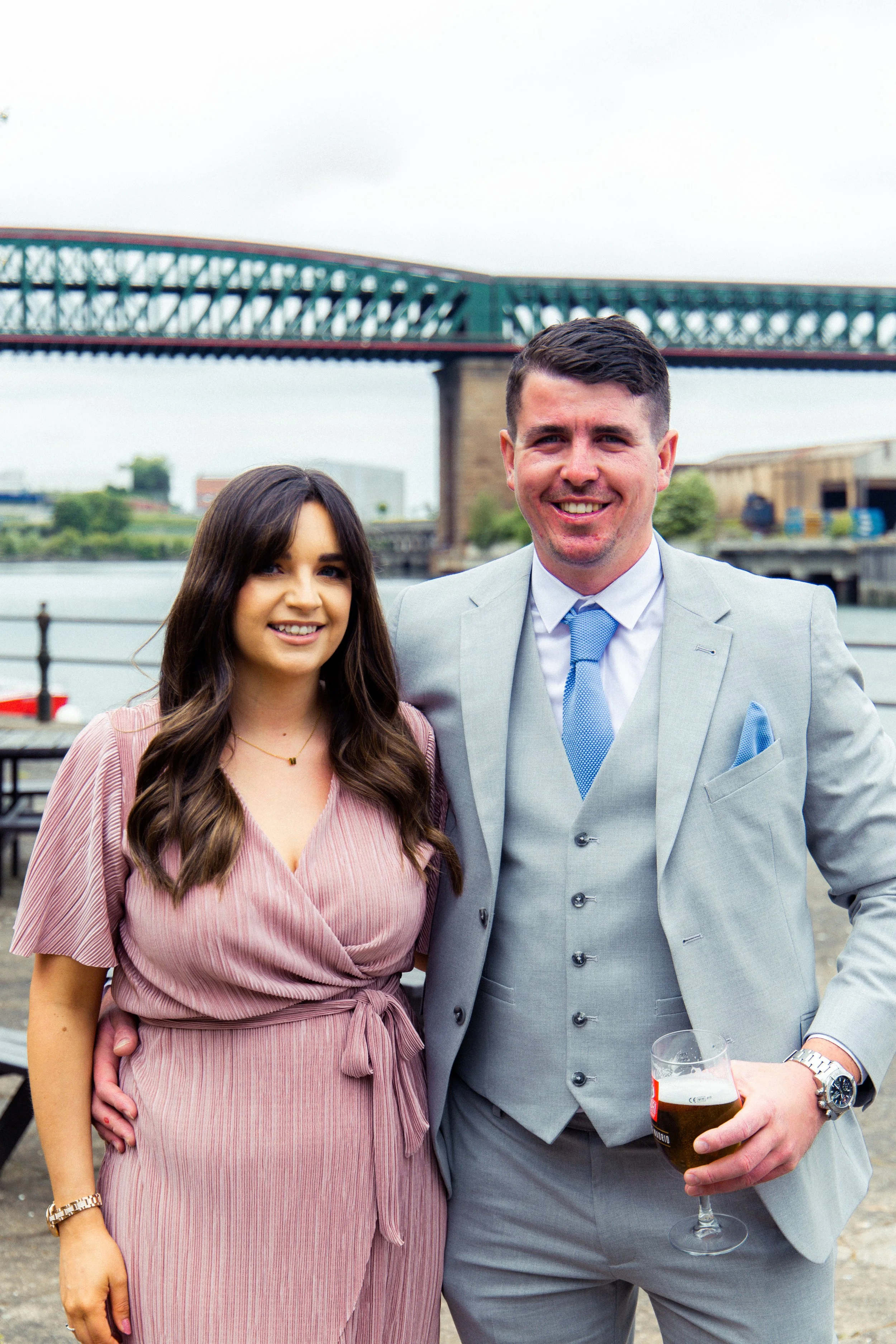 A man and woman standing outdoors near a body of water with a bridge in the background. The man is holding a glass of beer and is dressed in a light gray suit with a blue tie. The woman is wearing a pink dress and has long brown hair.
