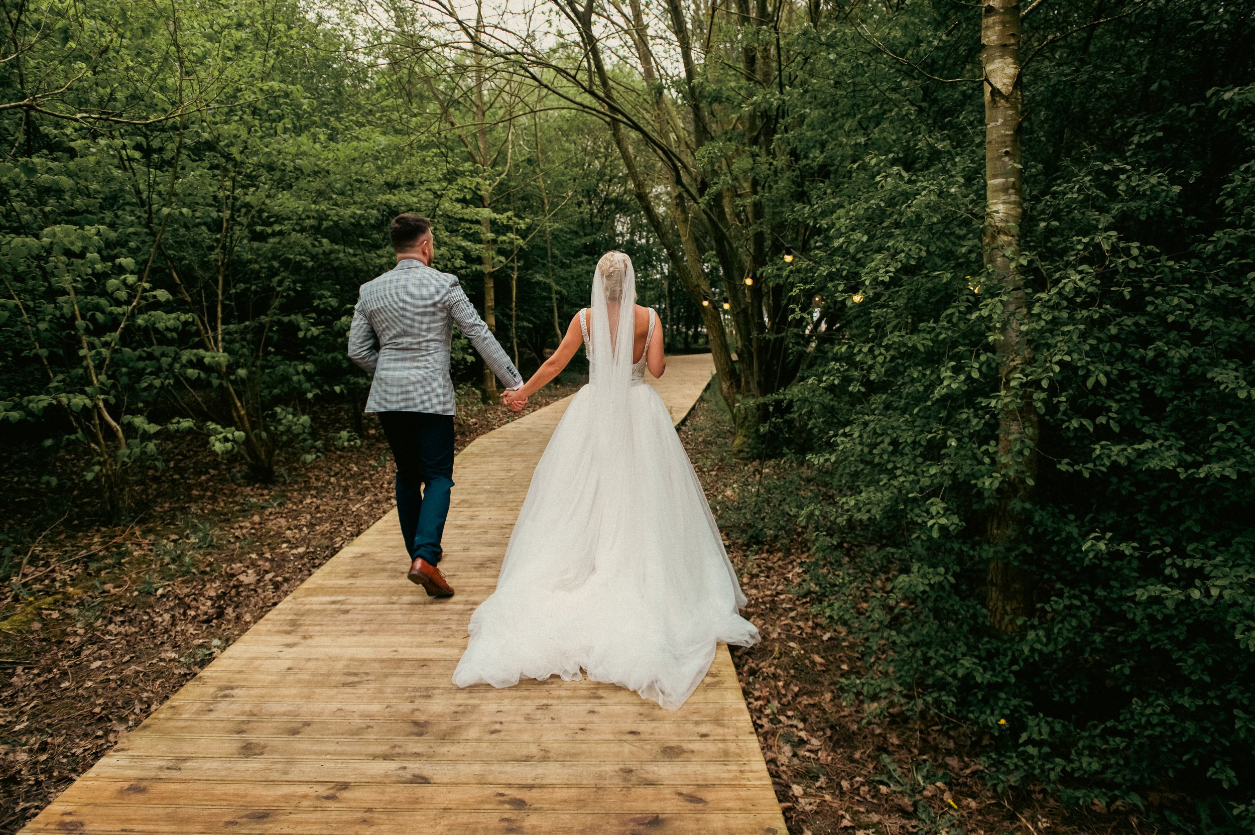 Bride and groom walking hand in hand on a wooden path through a lush green forest, with string lights hanging among the trees.