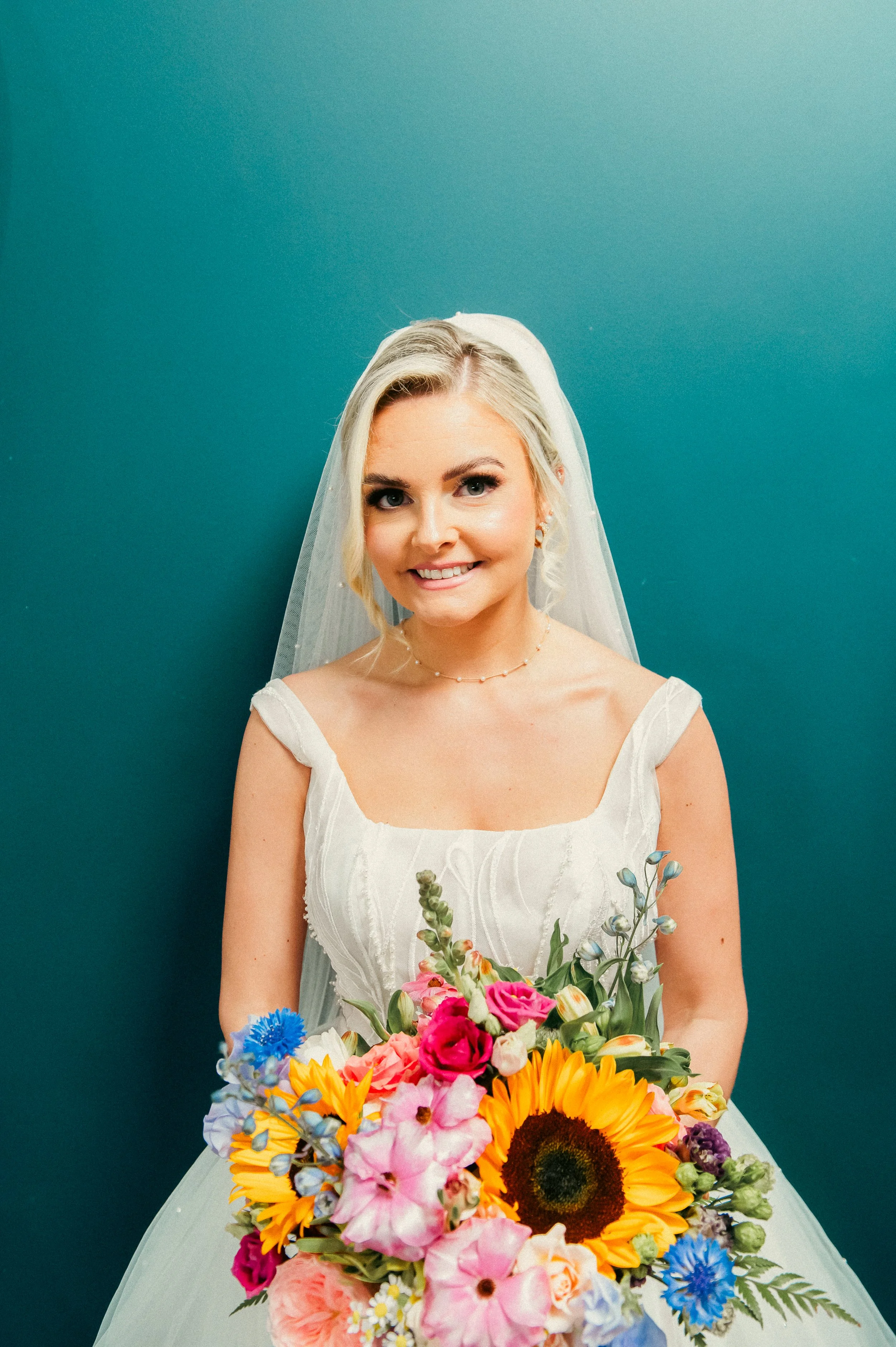 A smiling bride with blonde hair, wearing a white wedding dress and veil, holding a large bouquet of colorful flowers, standing against a teal background.