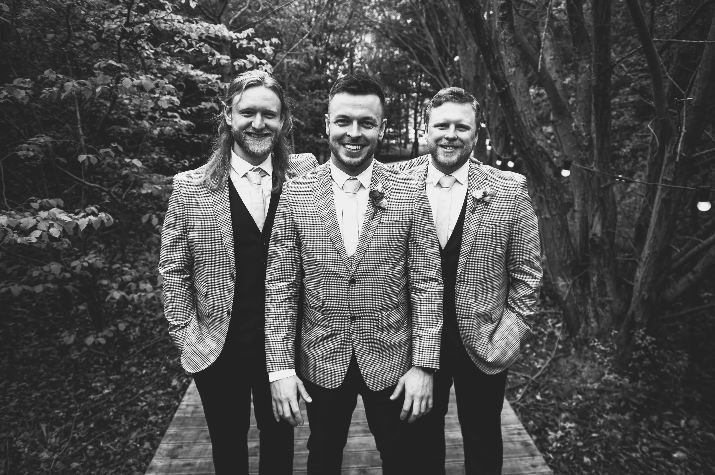 Three men in suits smiling for a photo outdoors on a wooden path, trees in the background, in black and white.
