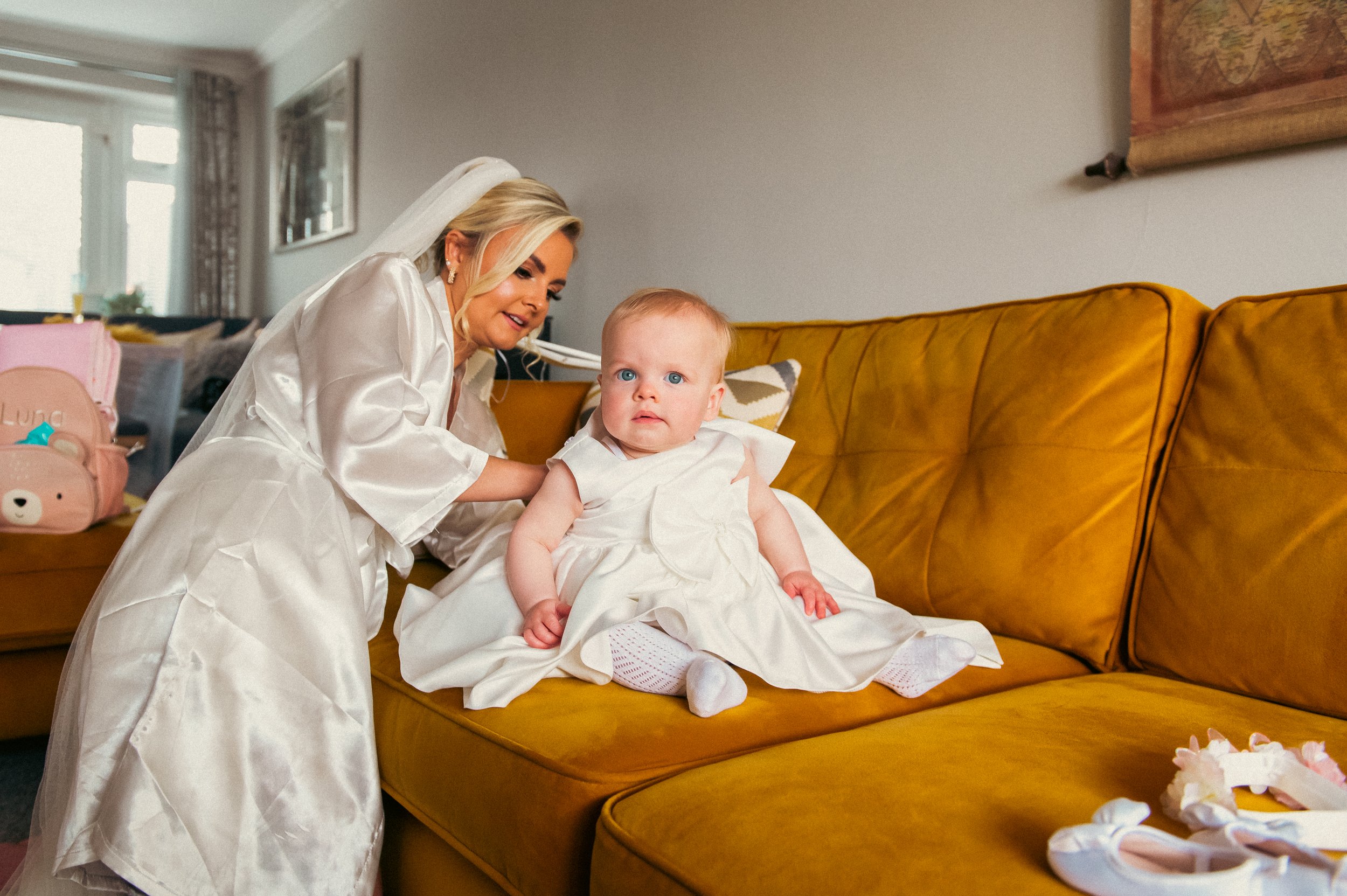 A woman in a white satin robe, wearing a veil, is leaning over a toddler girl sitting on a mustard-colored sofa, both dressed in white. The toddler has blue eyes and blonde hair, and is looking slightly upward with a curious expression. The backgroun
