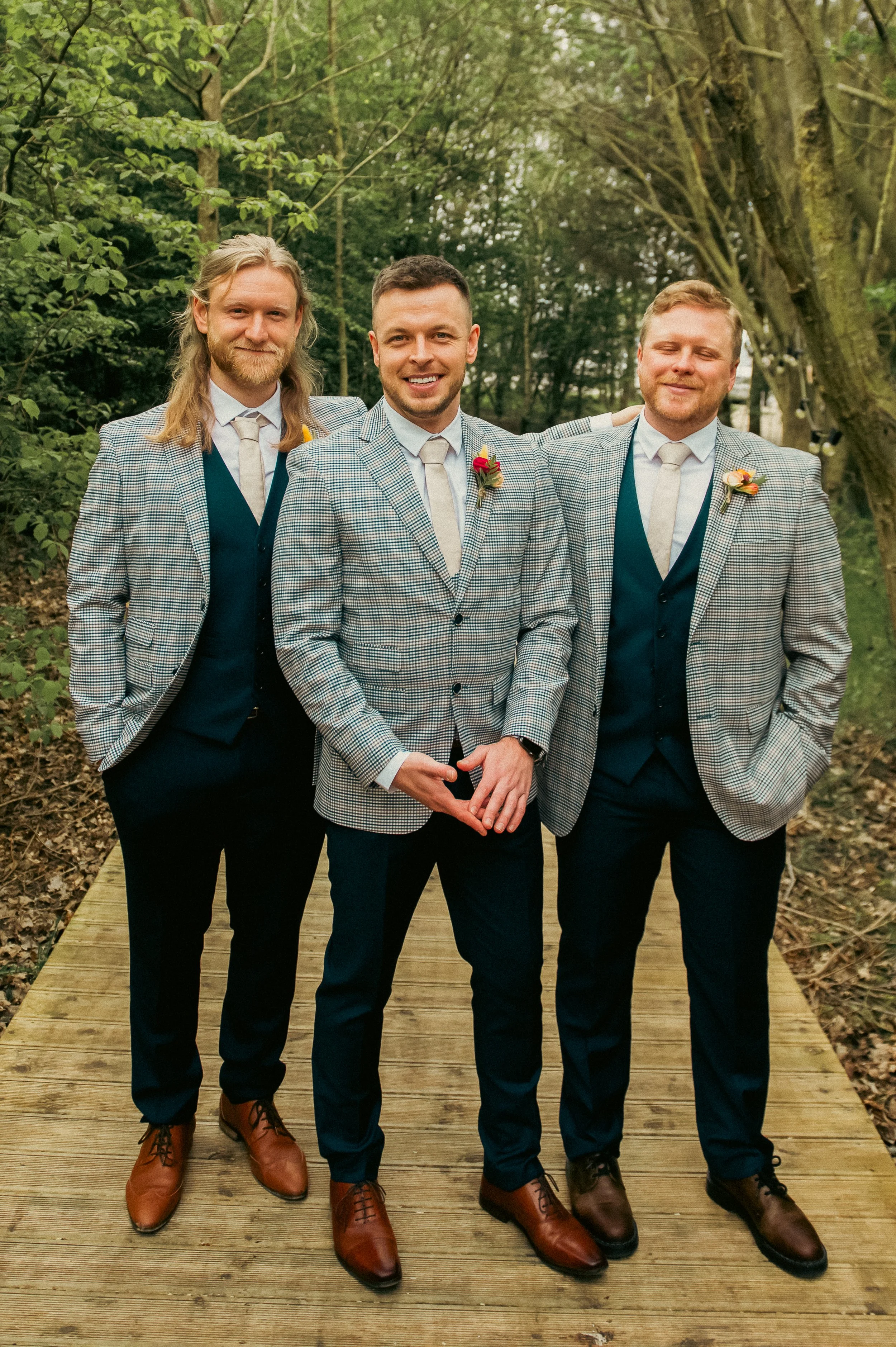 Three men dressed in matching suits and ties, standing on a wooden pathway outdoors in a wooded area, smiling for a photo.