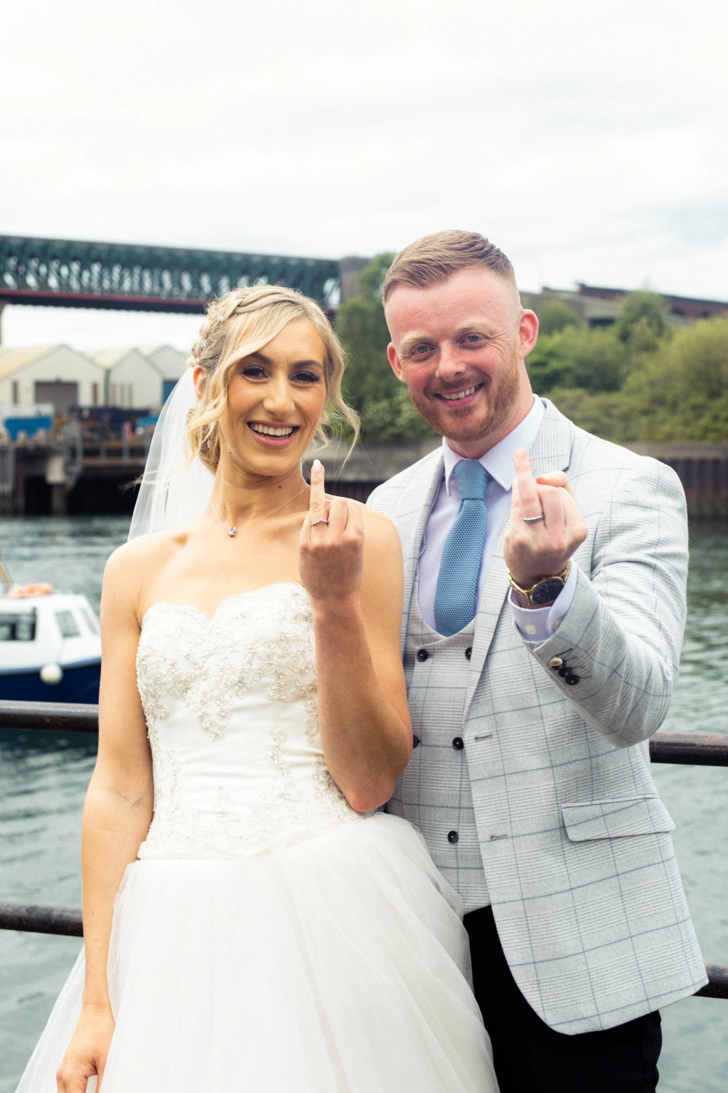 A happy bride and groom showing their wedding rings and middle fingers during their wedding photos near a river with boats, bridge, and buildings in the background.