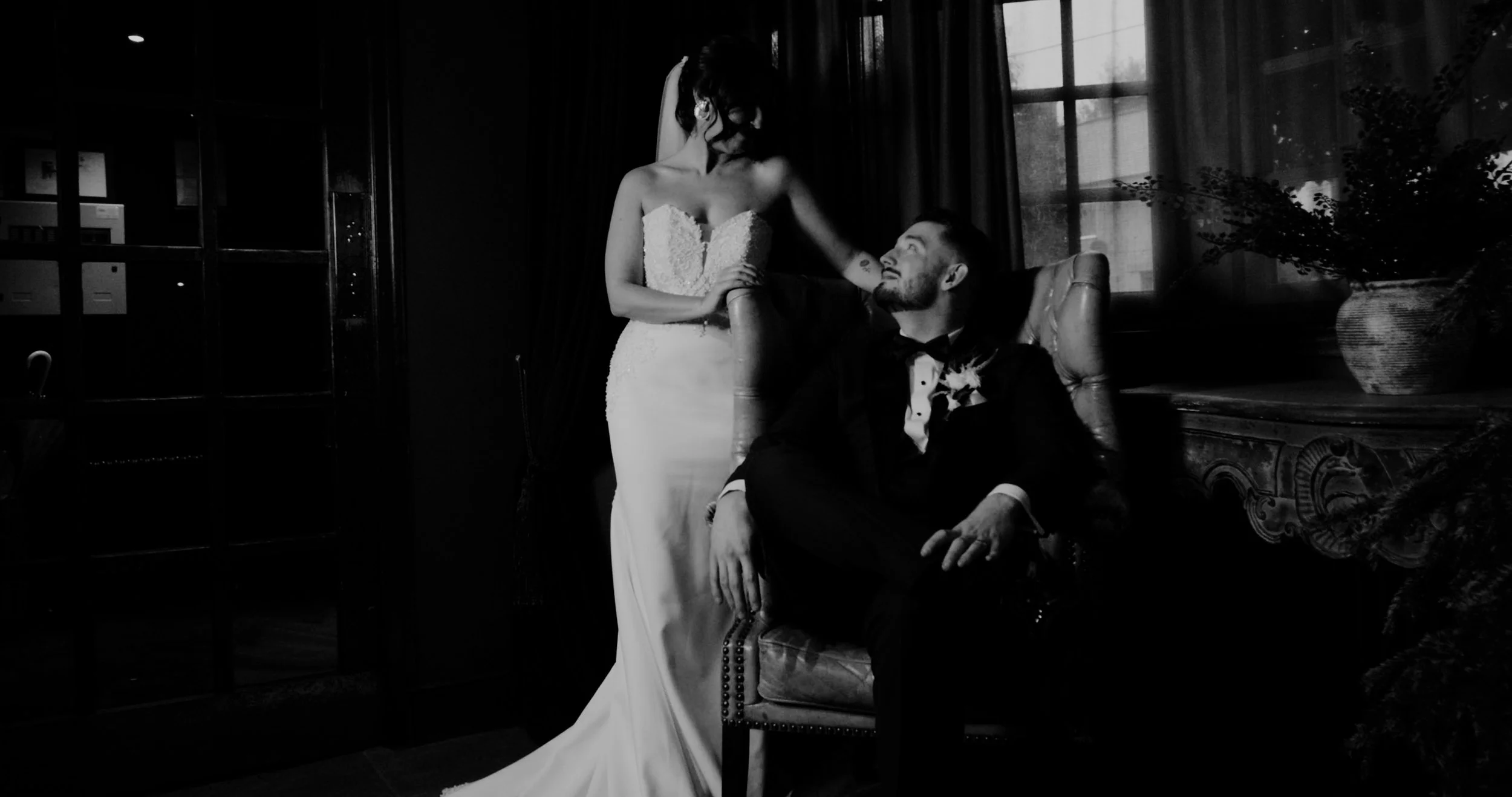 A black and white photo of a bride and groom indoors. The bride is standing, wearing a strapless wedding gown, while the groom is sitting on a chair, wearing a tuxedo. The bride is touching the groom's shoulder and looking at him.