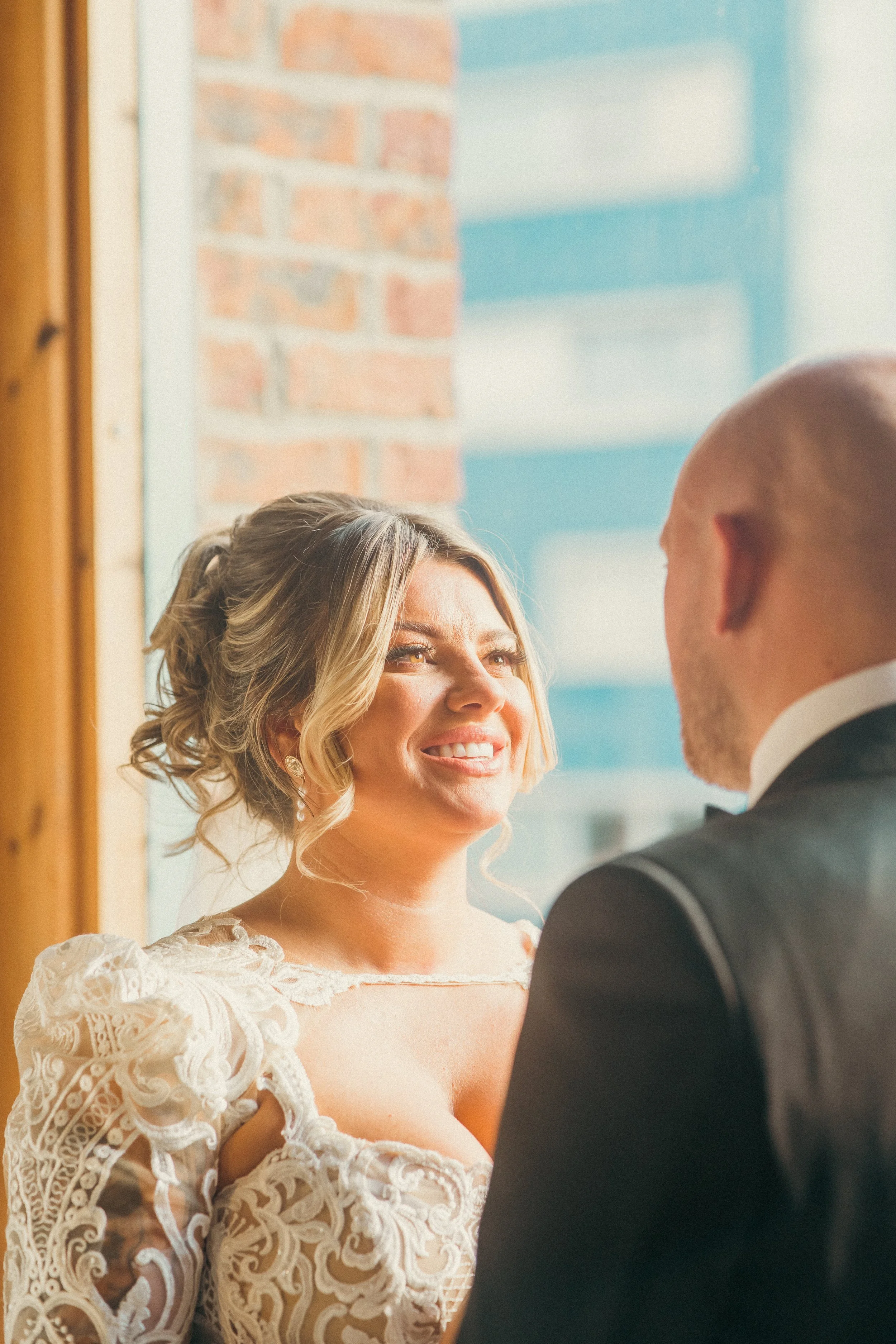 A bride and groom during their wedding ceremony, facing each other by a window with a brick wall and building in the background.