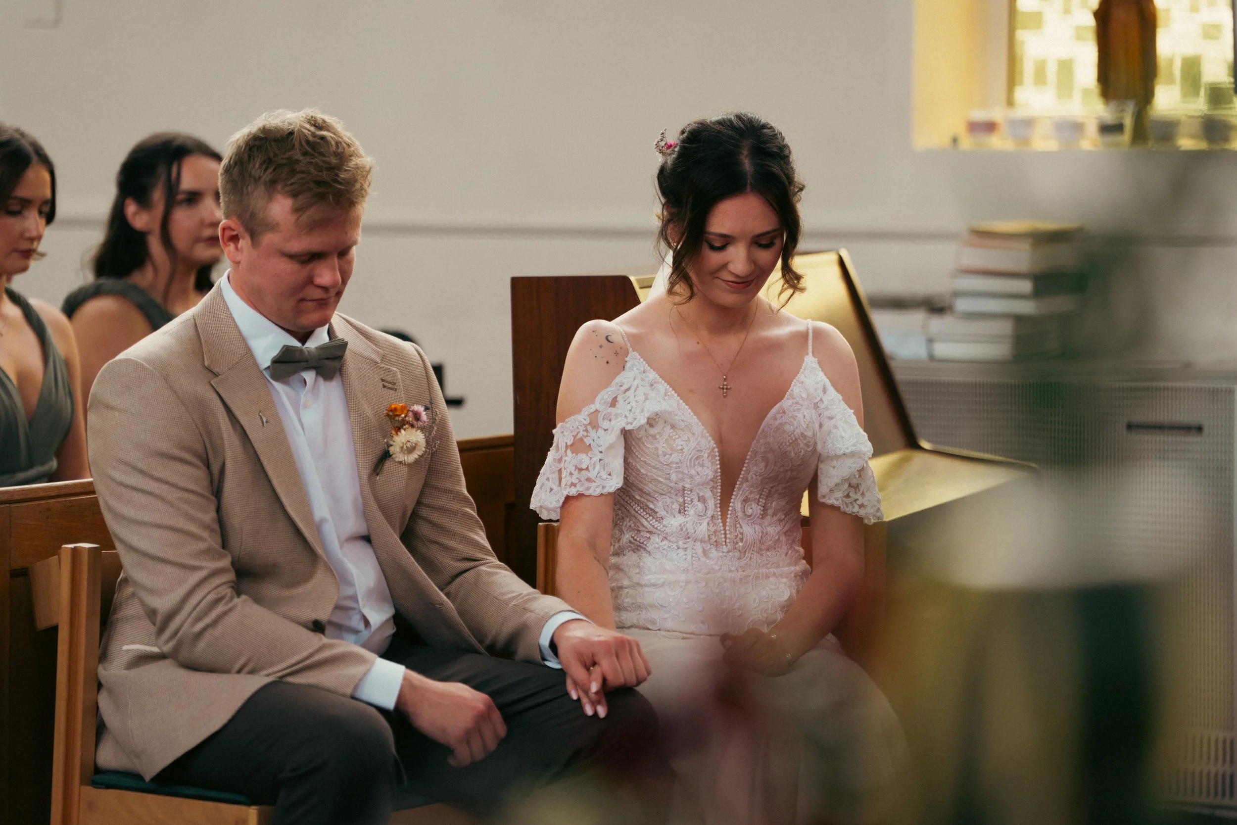 A bride and groom sitting side by side during a wedding ceremony, with the bride holding the groom's hand and both looking down, surrounded by guests in a church or chapel setting.