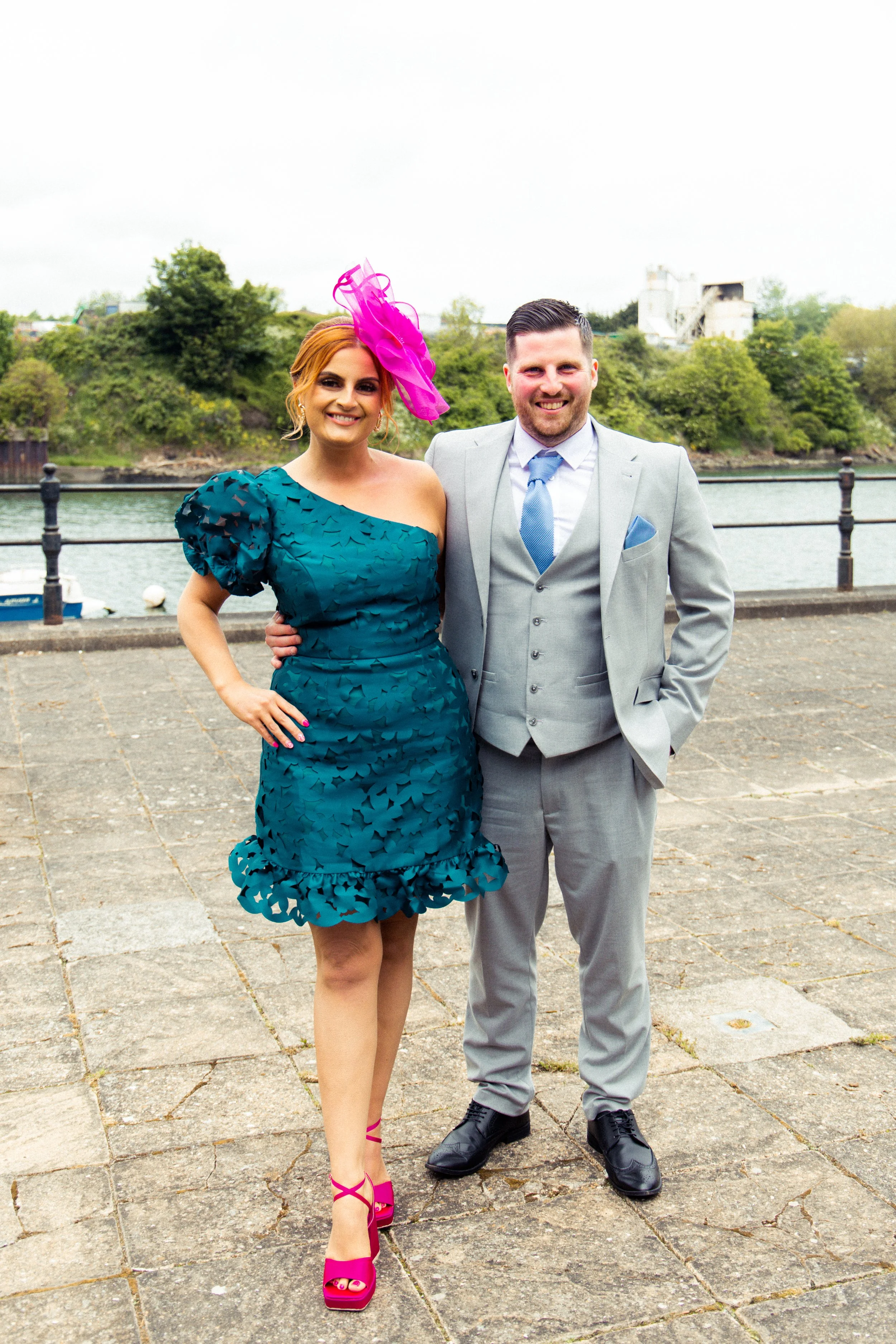 A woman in a teal dress with ruffle details and a pink fascinator, standing next to a man in a light gray suit and blue tie, outdoors near a body of water with greenery in the background.