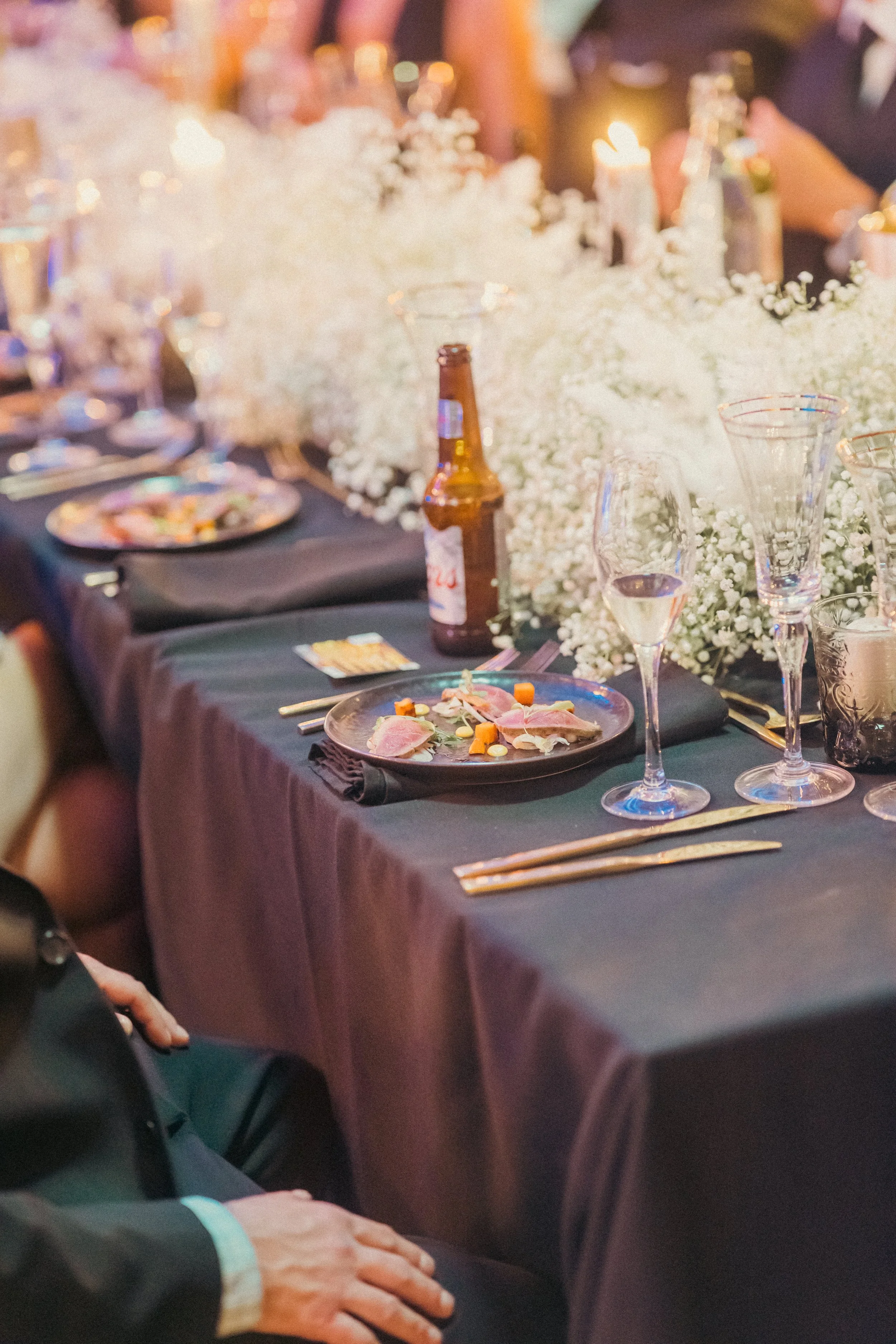 A formal dining table set with black tablecloth and elegant glassware, featuring plates of food, a bottle of beer, white floral centerpieces, candles, and place settings.