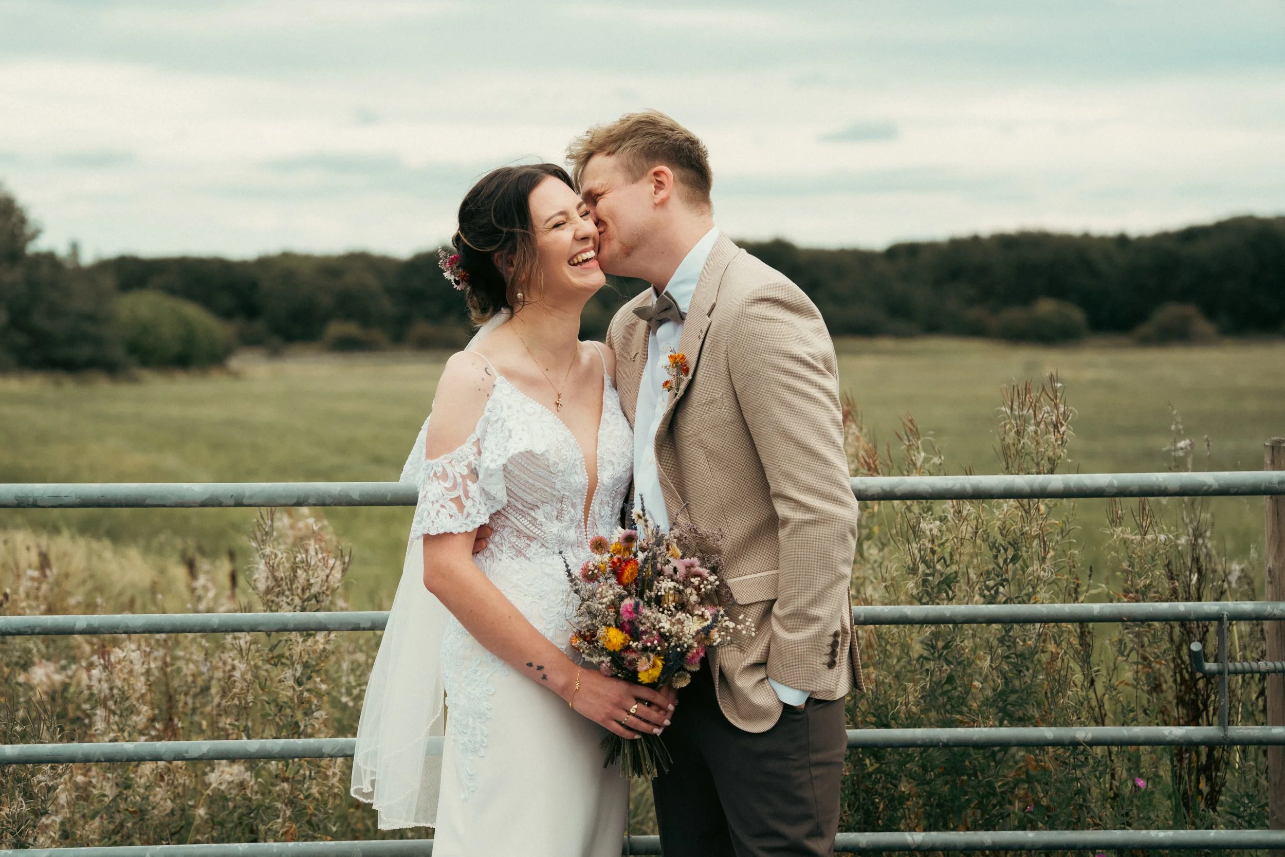 A bride and groom sharing a kiss on their wedding day outdoors, with a pasture and cloudy sky in the background.