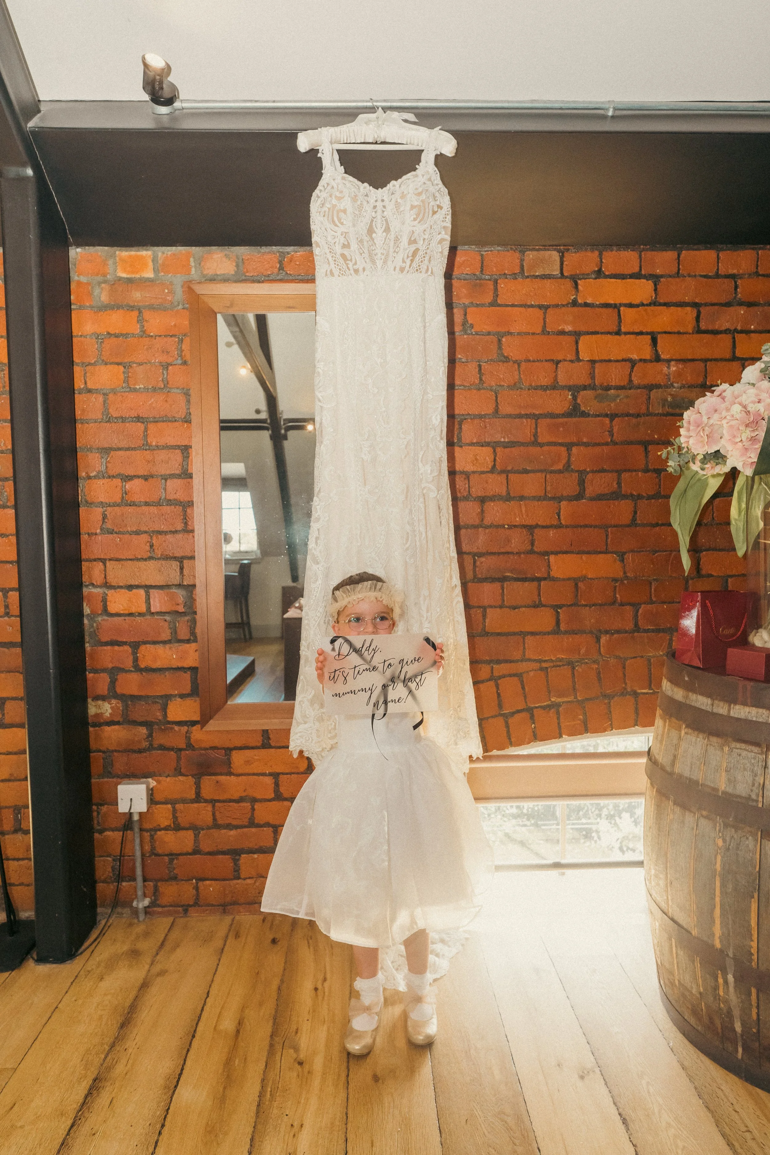 A young girl dressed in a white dress and shoes stands in front of a hanging wedding dress, holding a sign that reads 'Daddy, it's time to give mommy our last name!'.