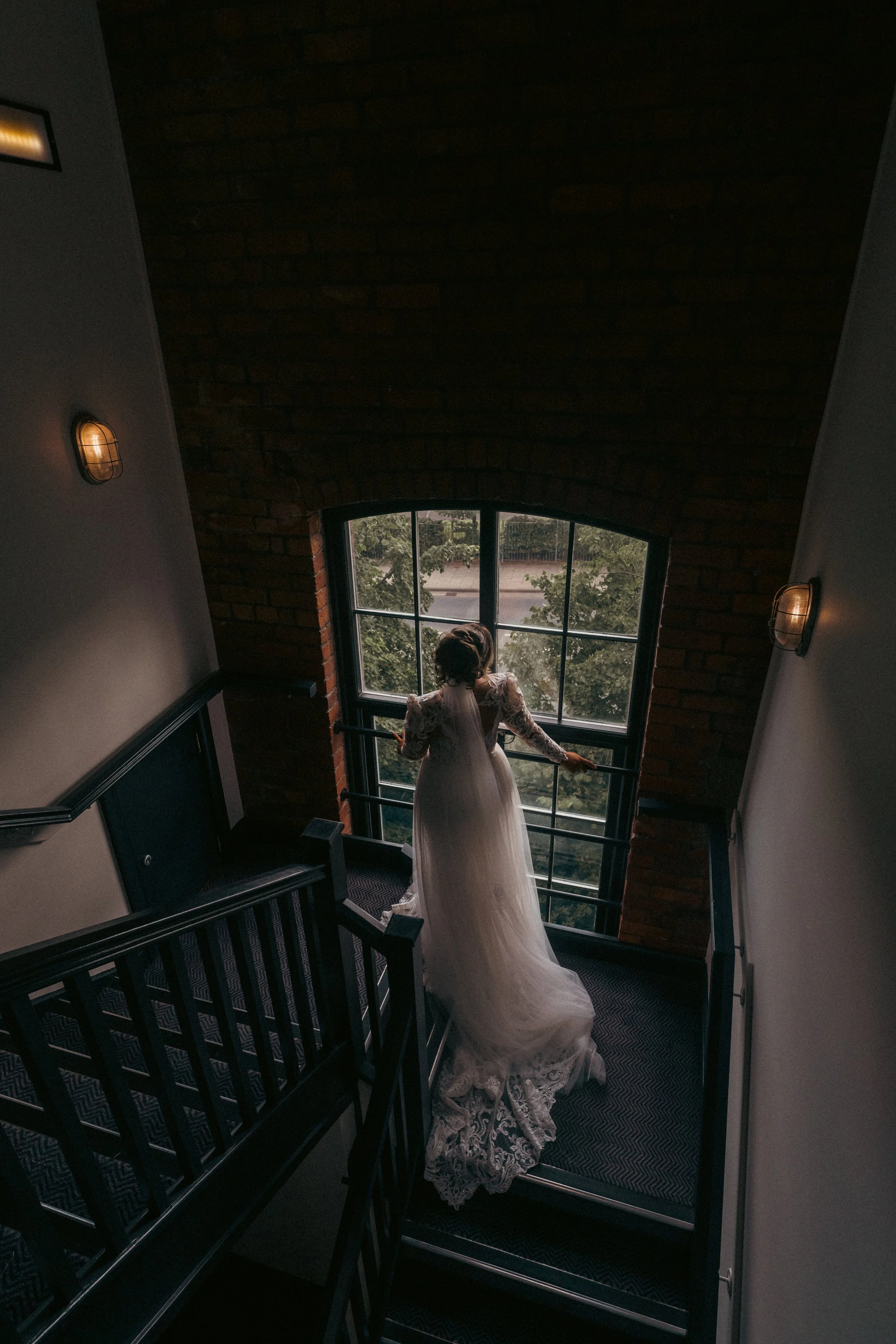 A woman in a lace wedding dress standing on a staircase, looking out a large window with trees outside, in a dimly lit interior with brick wall and wall-mounted lights.