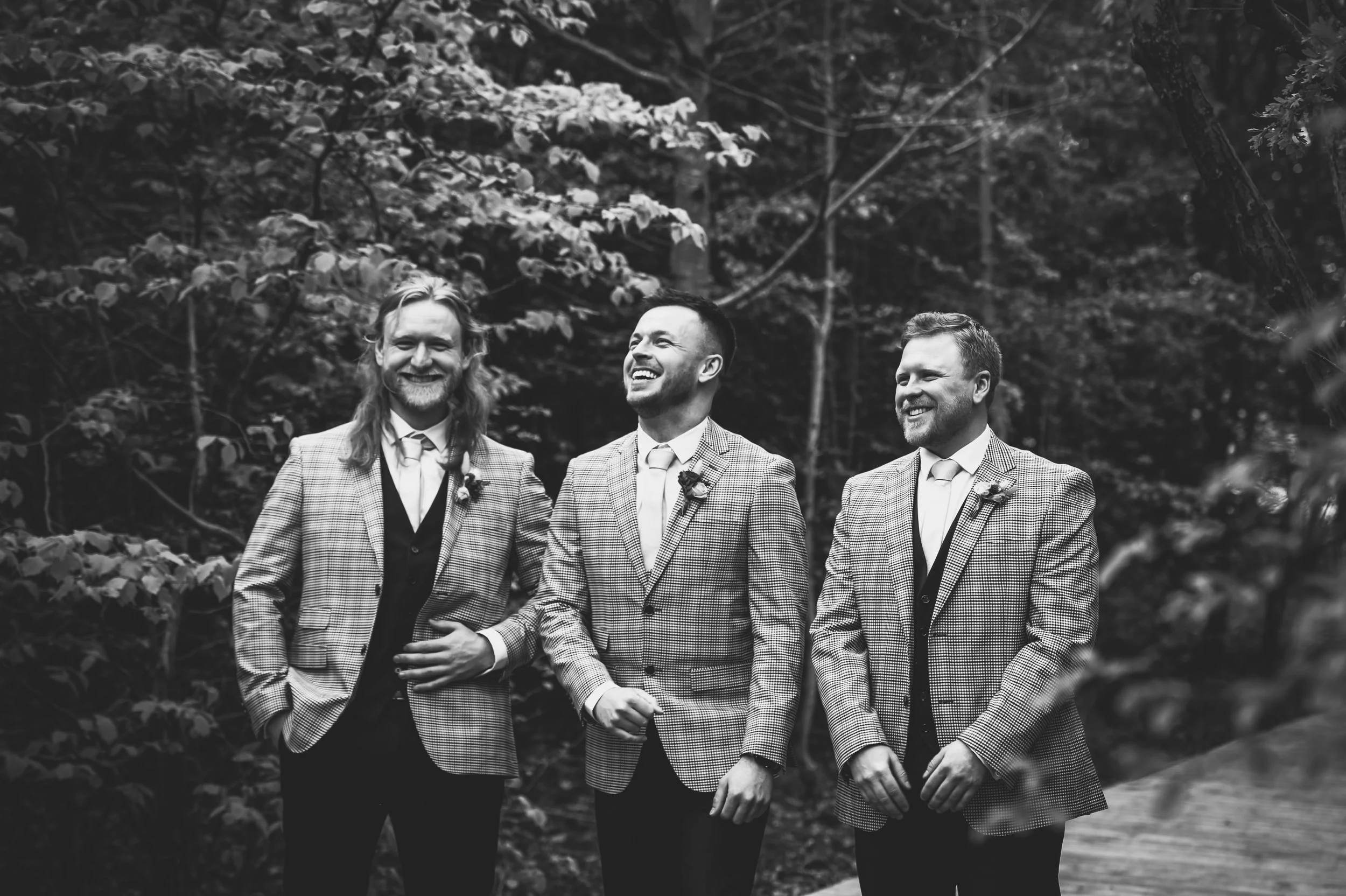 Three men in plaid suits and vests, smiling and standing outdoors in a wooded area.