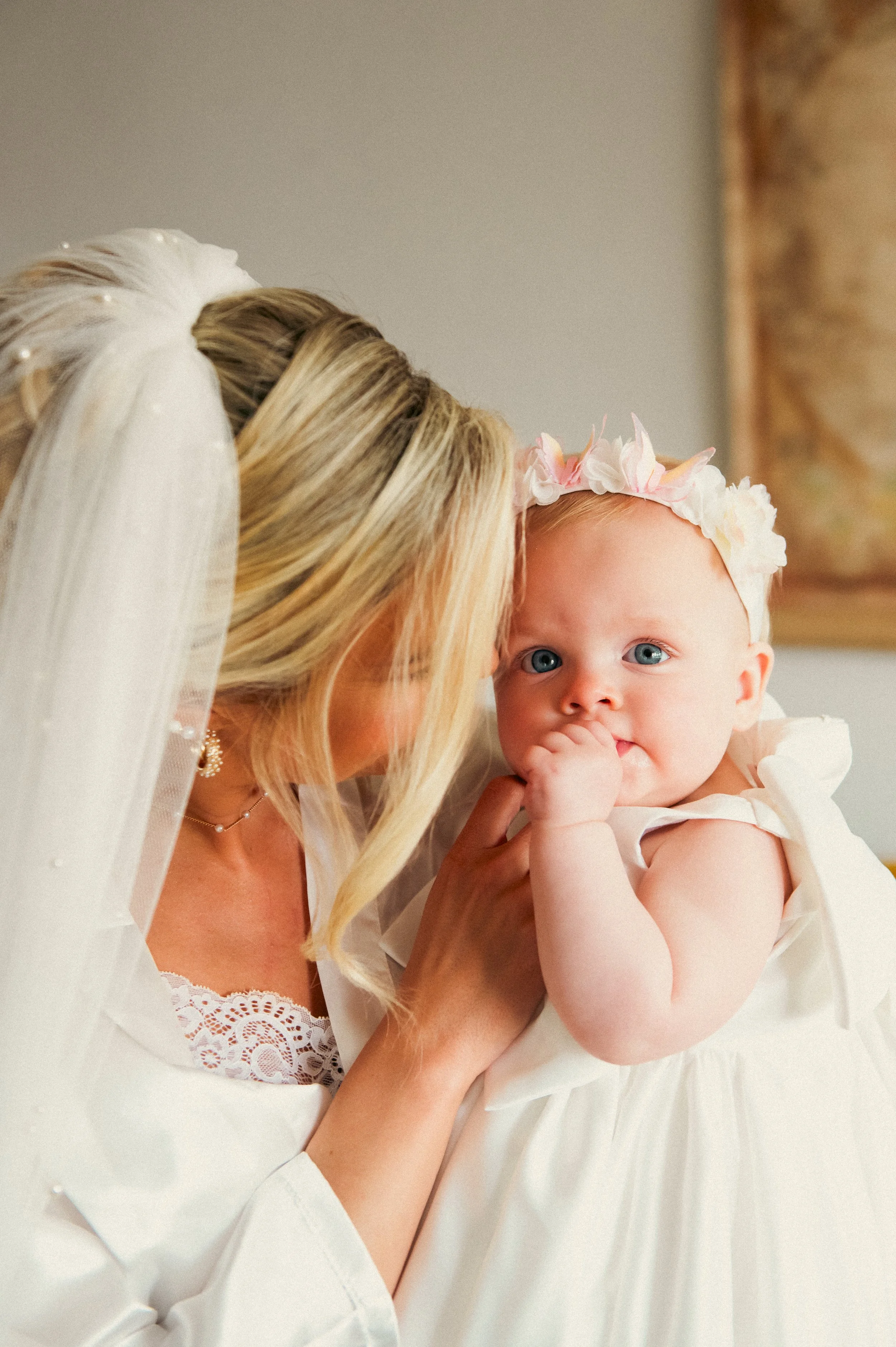 A bride holding a baby girl, both dressed in white, with the baby wearing a pink flower crown and sucking her thumb.
