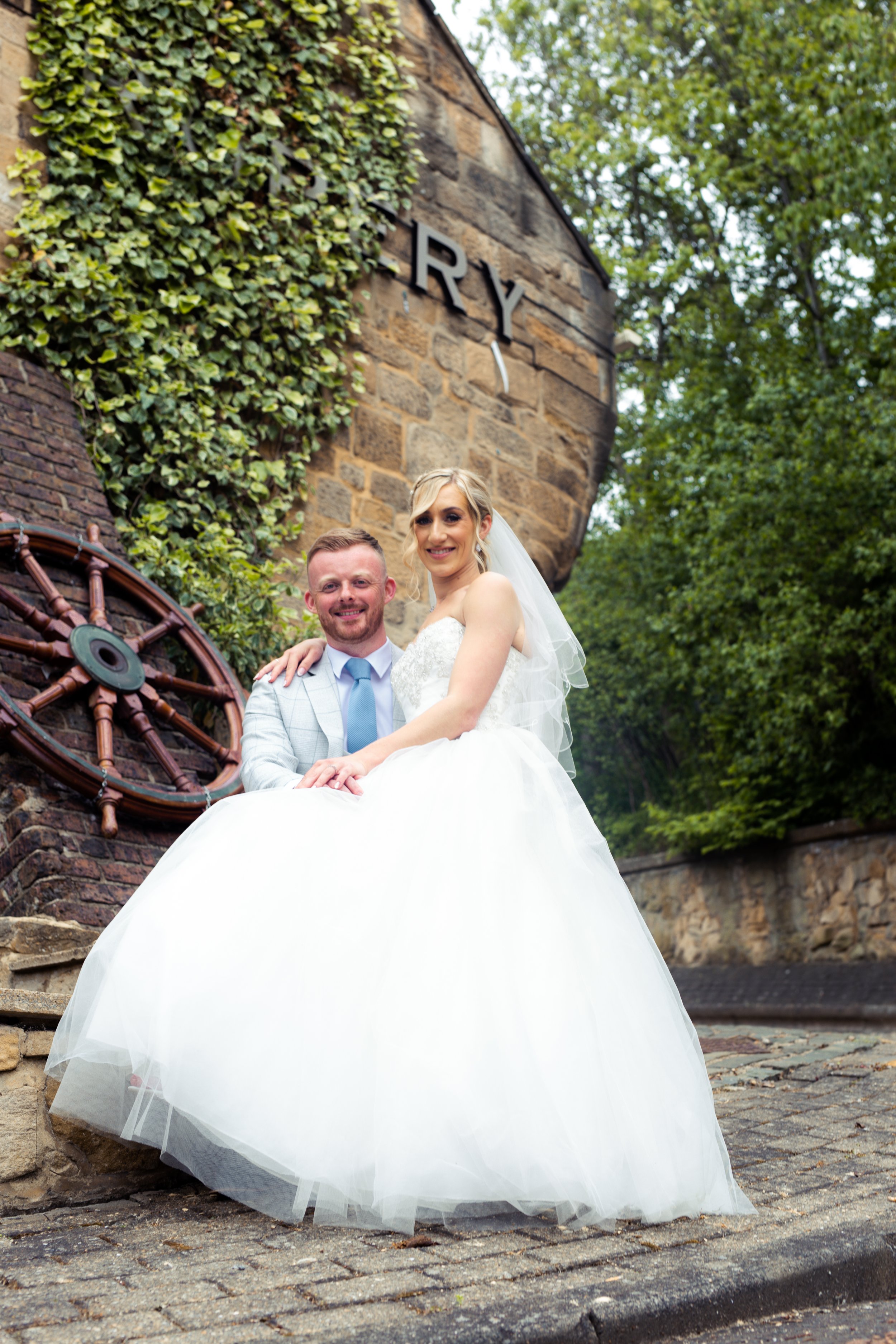 A bride and groom sitting on a stone ledge outdoors, smiling, with a rustic brick wall, a nautical wheel decoration, and green trees in the background.