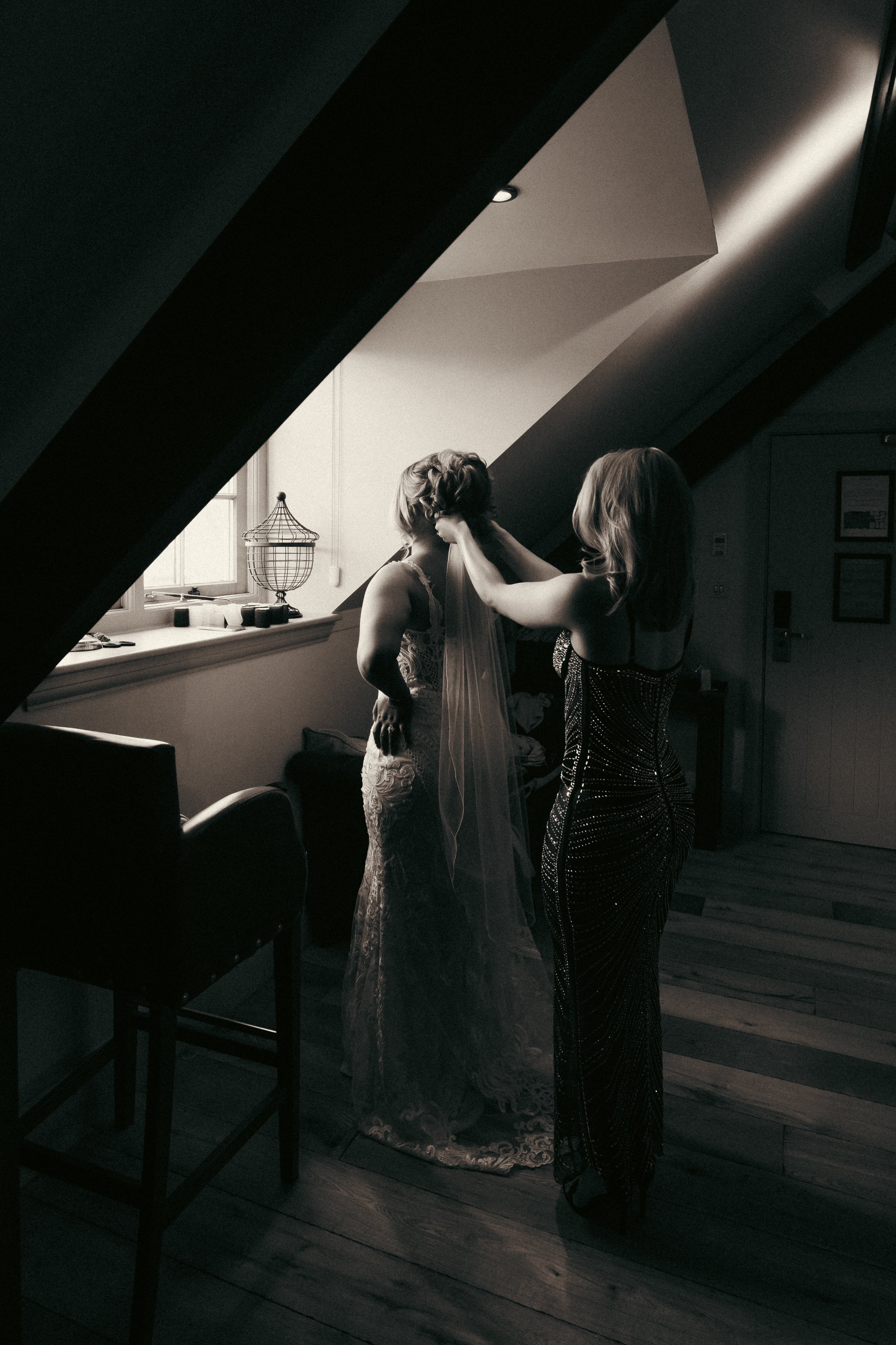 Two women are in a room with sloped ceilings. One woman is helping the other with a veil, and the woman receiving help is wearing a lace wedding dress. The room has a window with a small lamp and decorative items on the window sill.