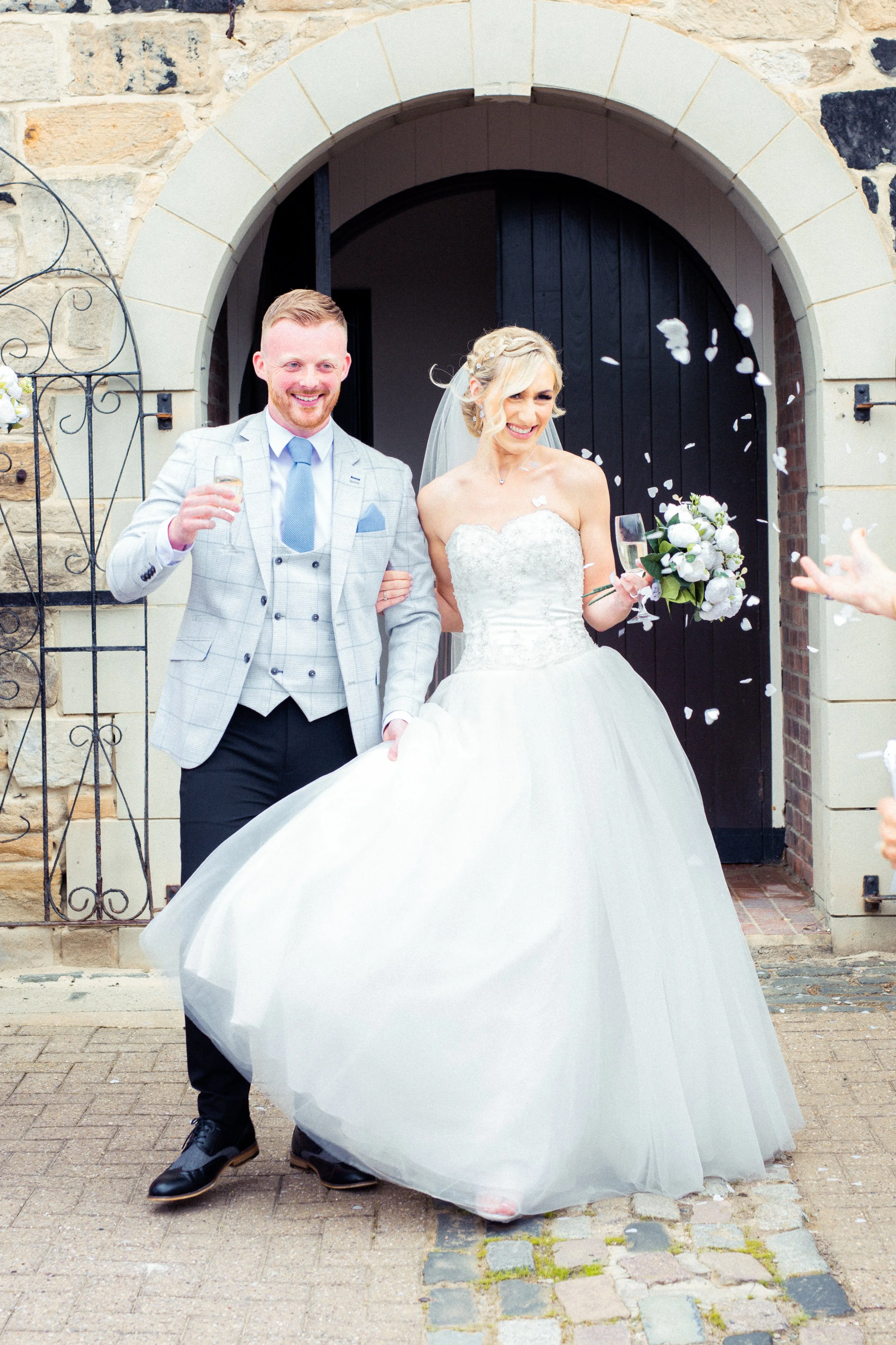 Bride and groom smiling as they walk out of a church, being showered with flower petals, holding champagne glasses, bride holding a bouquet, man in tuxedo, woman in wedding dress.
