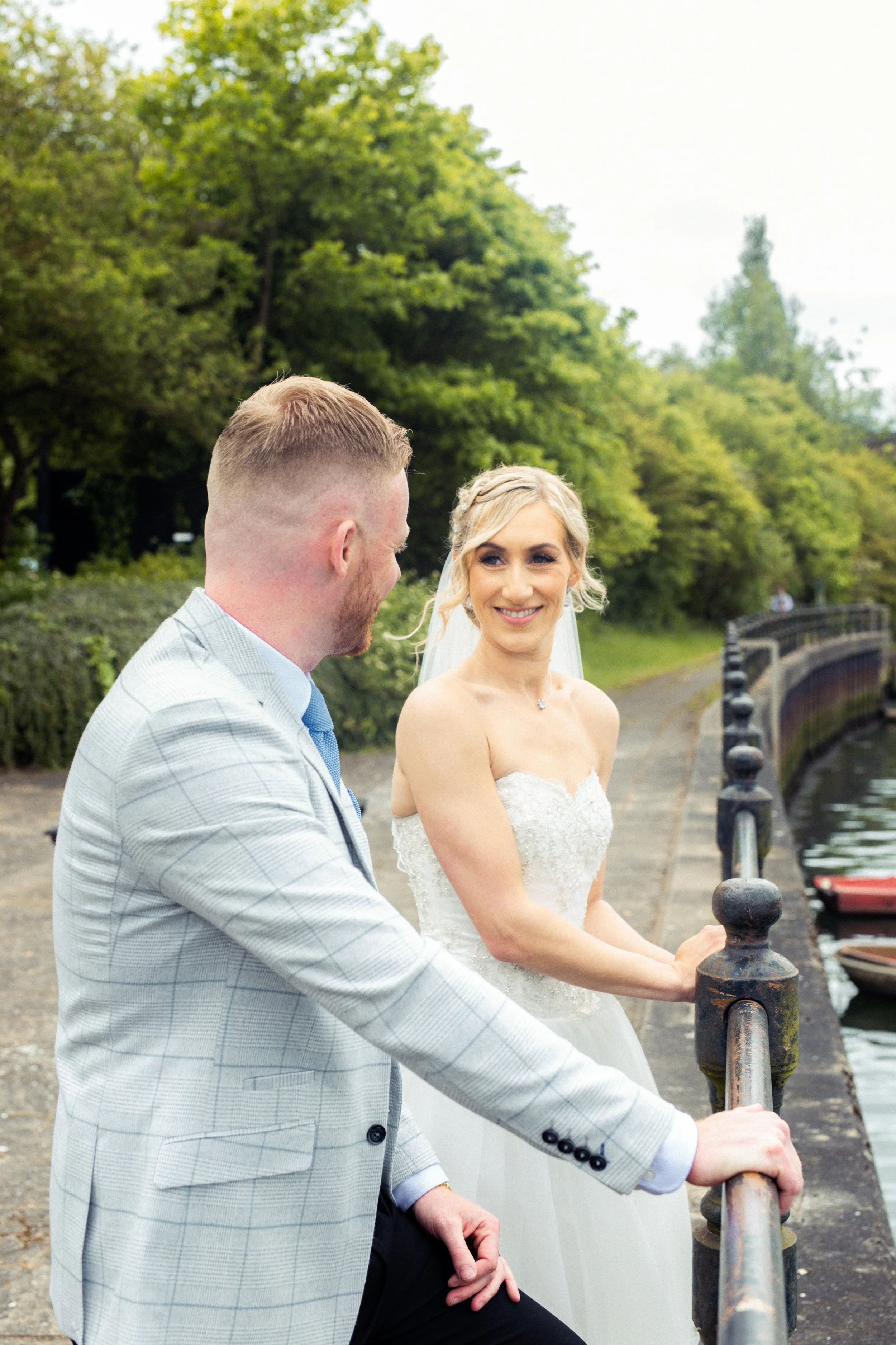 A bride and groom standing by a river, holding hands and smiling at each other, with trees and a pathway in the background.