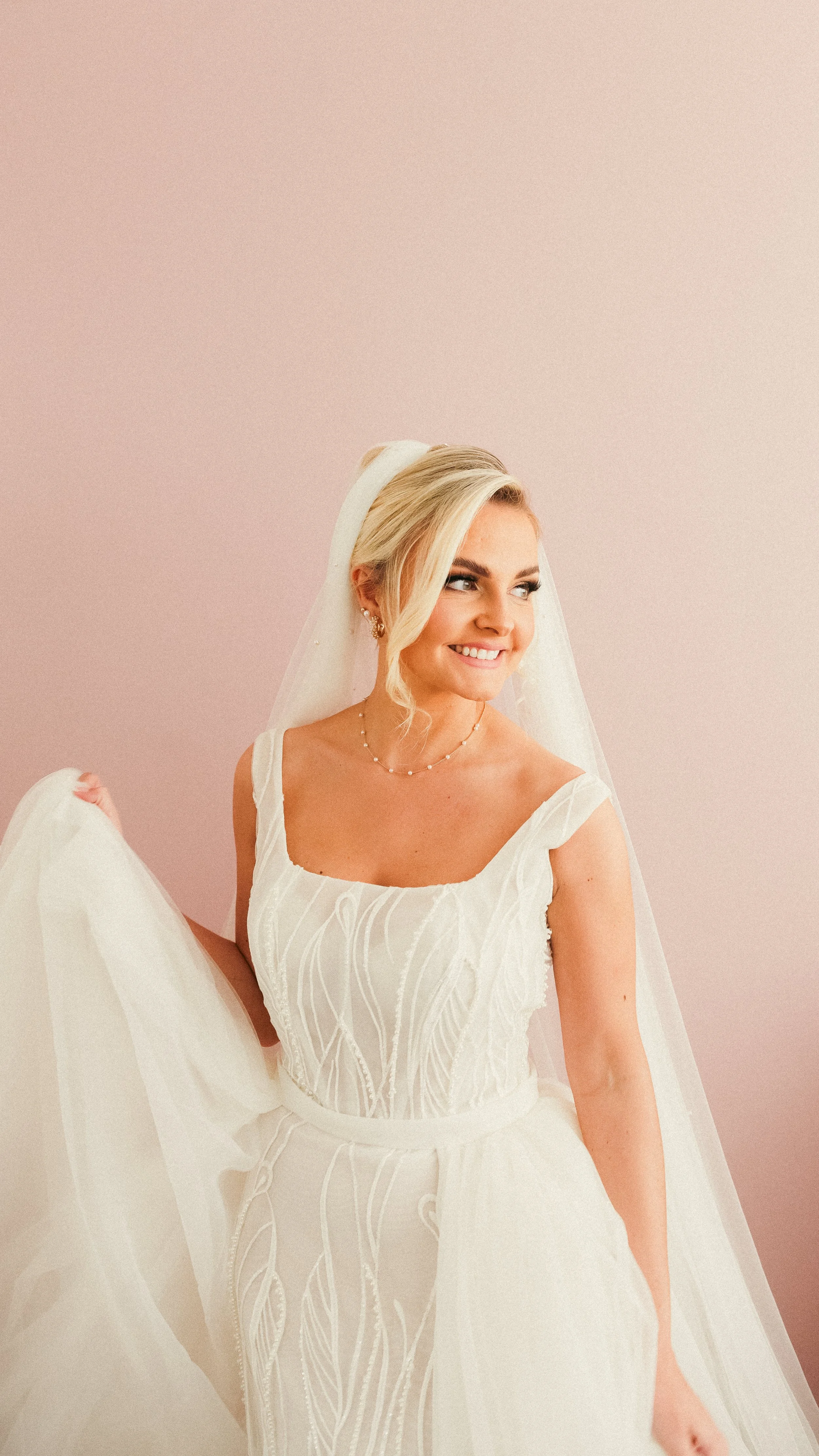 A bride wearing a white wedding dress with lace details, holding the skirt of her dress, standing against a light pink background, smiling and looking to her right.
