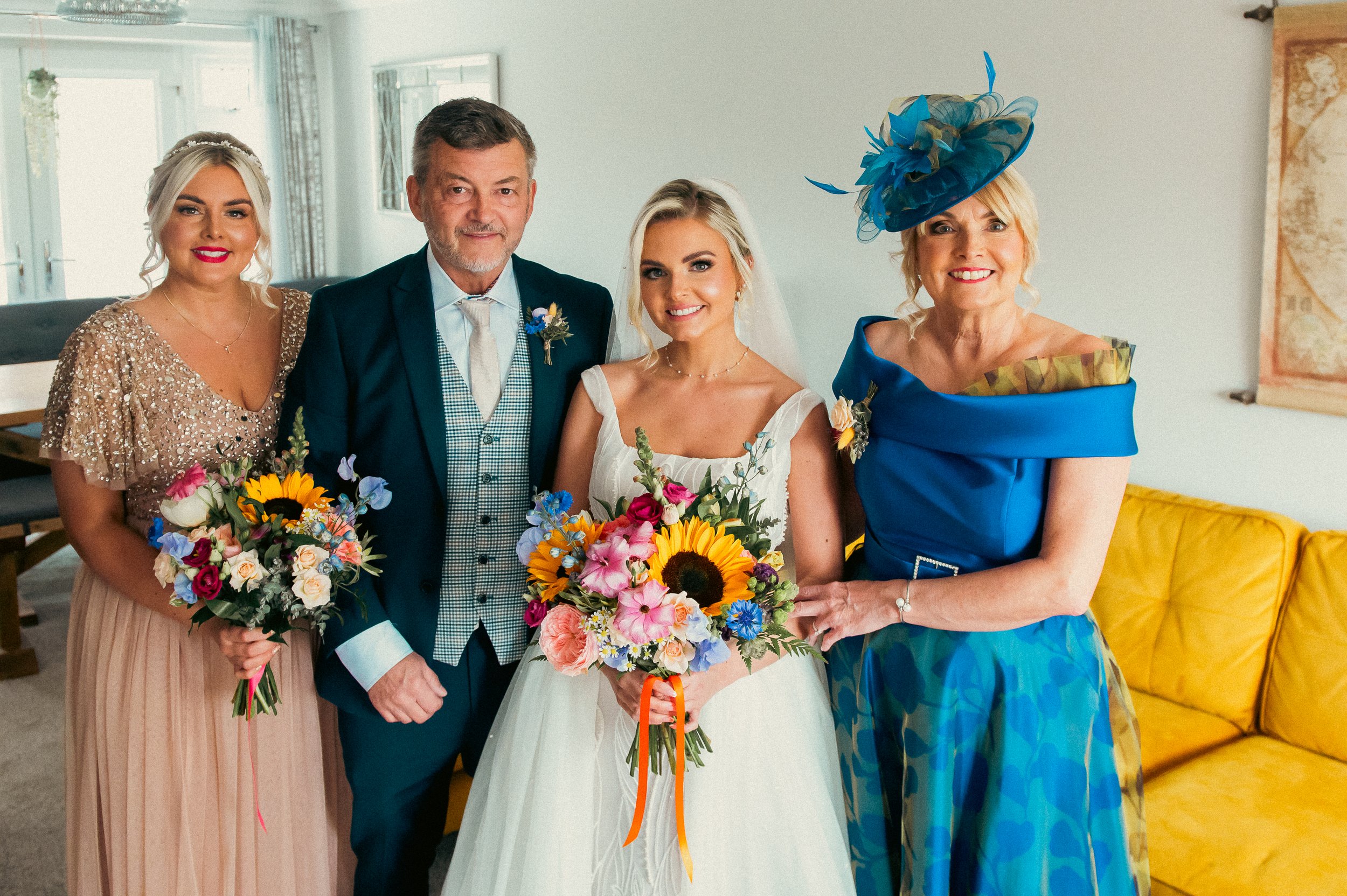 Group of five people at a wedding, including the bride in a white dress holding a colorful bouquet, standing between a man in a blue suit and a woman in a vibrant blue dress and hat, with another woman in a beige dress holding a bouquet.
