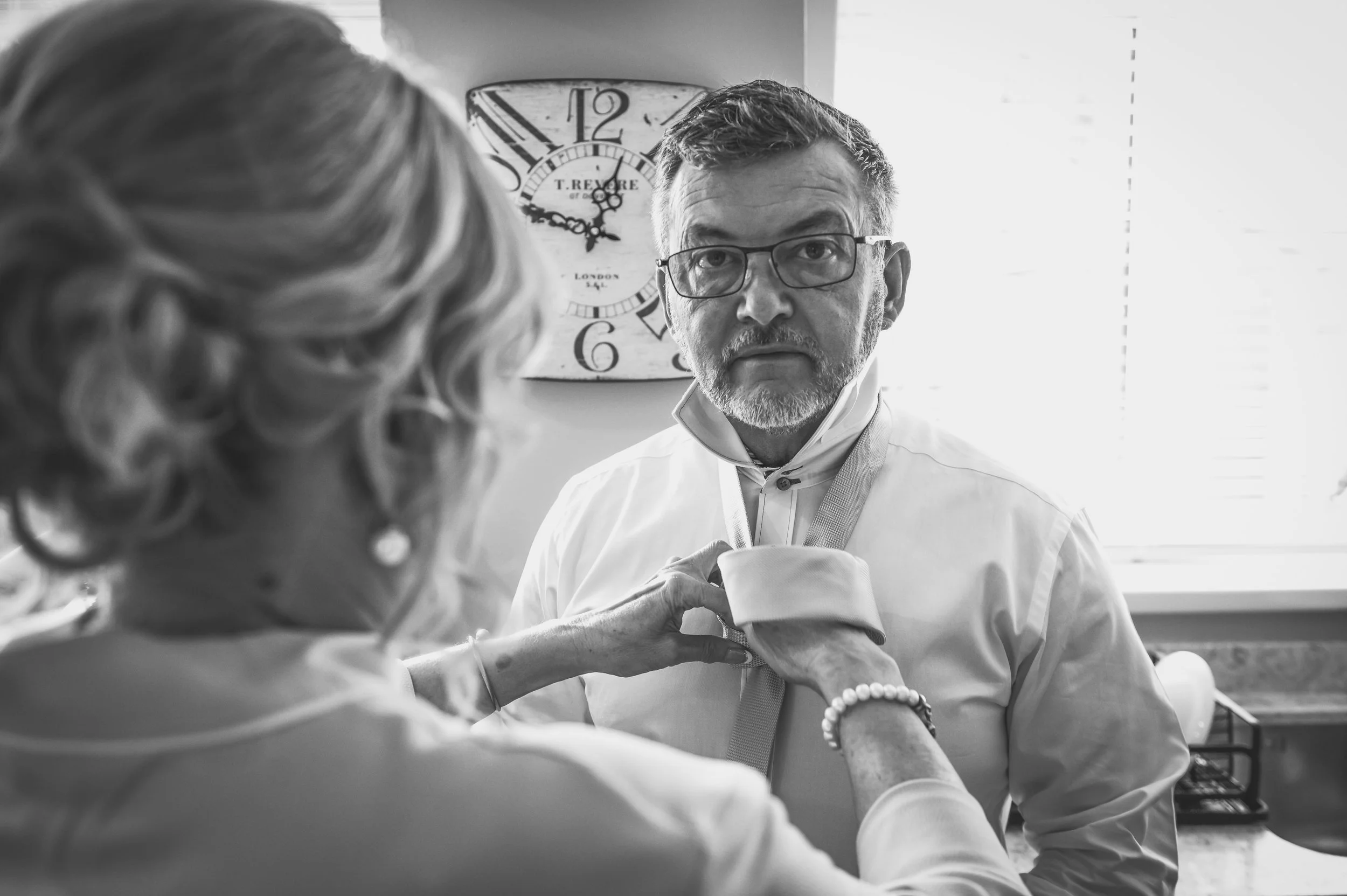 An older woman adjusting the collar of a man wearing glasses in a kitchen setting, with a wall clock in the background.