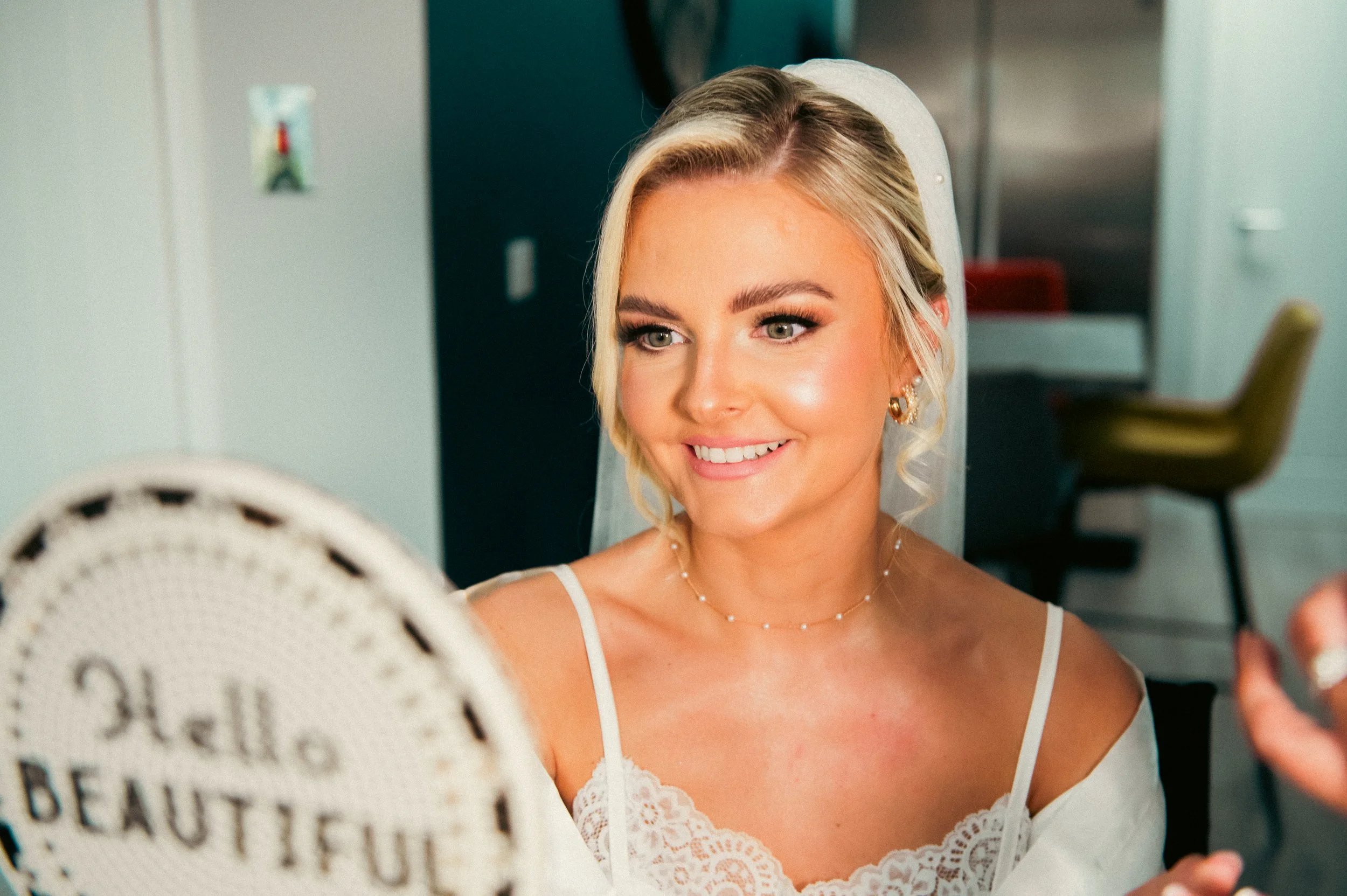 Bride with blonde hair and earrings smiling while looking at a mirror, wearing a white lace dress and veil, in a room with green and black walls.
