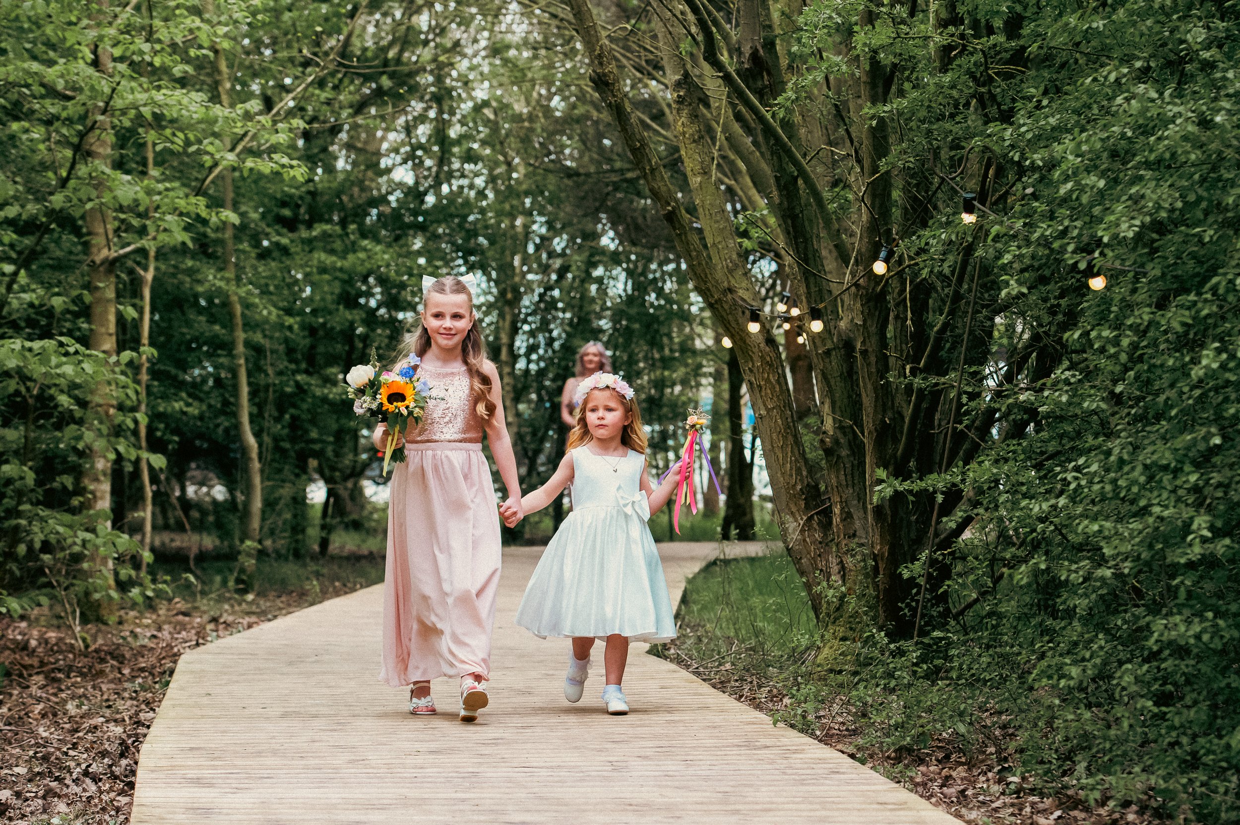 Two young girls dressed in fancy dresses walking hand-in-hand along a wooden pathway in a forest with string lights hanging from trees.