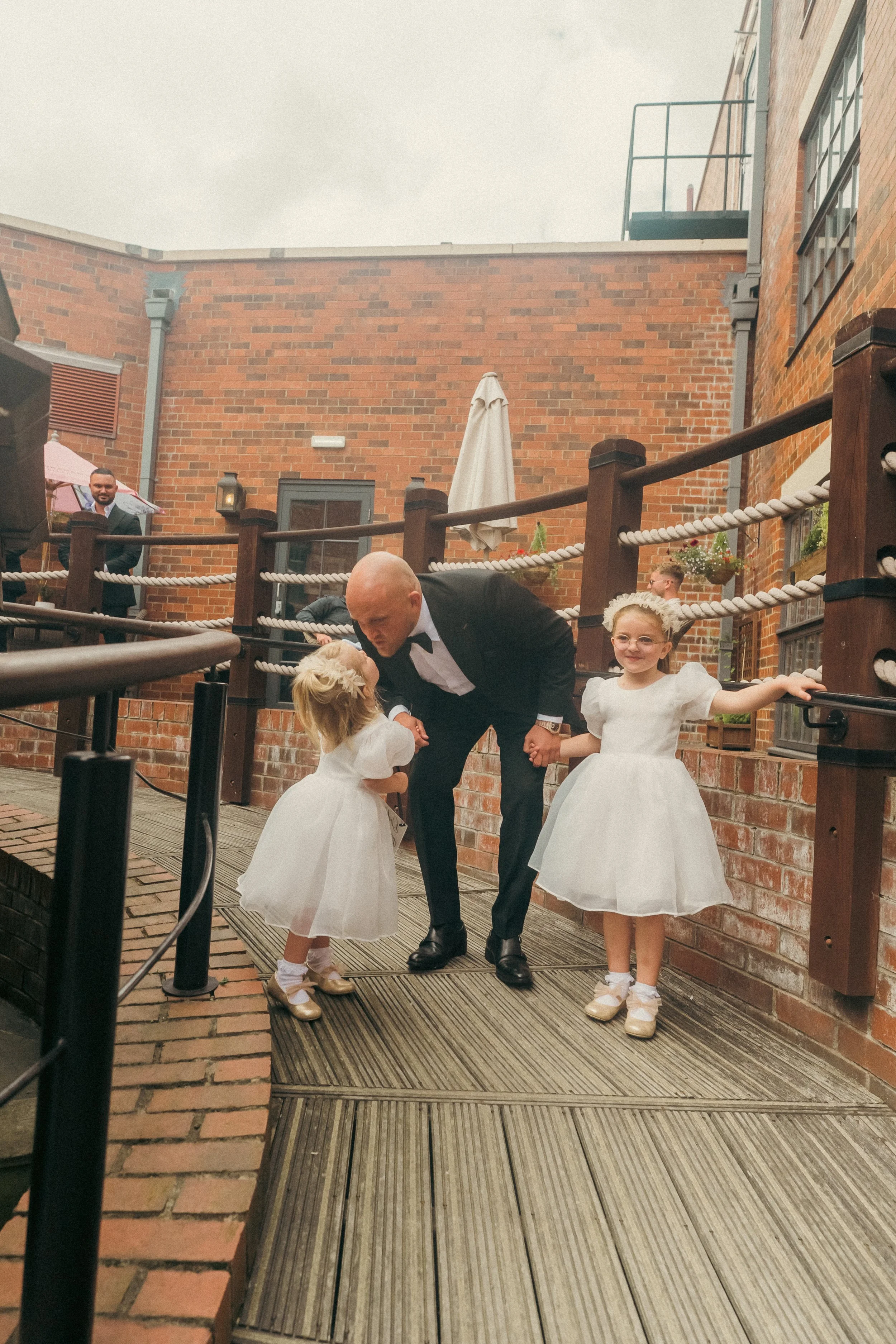 A man in a tuxedo kissing two young girls in white dresses on an outdoor wooden deck surrounded by brick walls and safety ropes, with guests in the background.