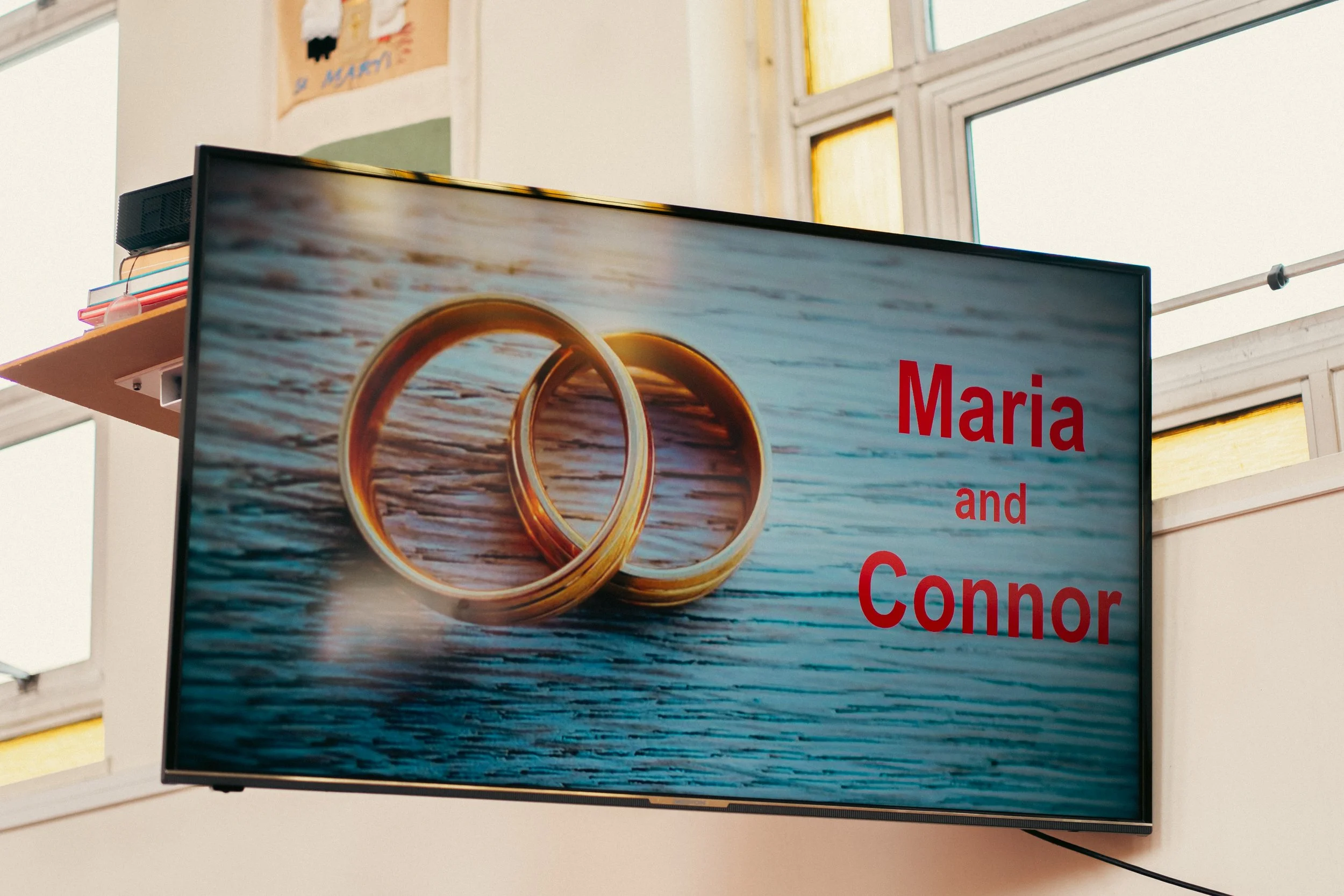 A digital sign showing two gold wedding rings on a wooden surface with the text "Maria and Connor" in red.