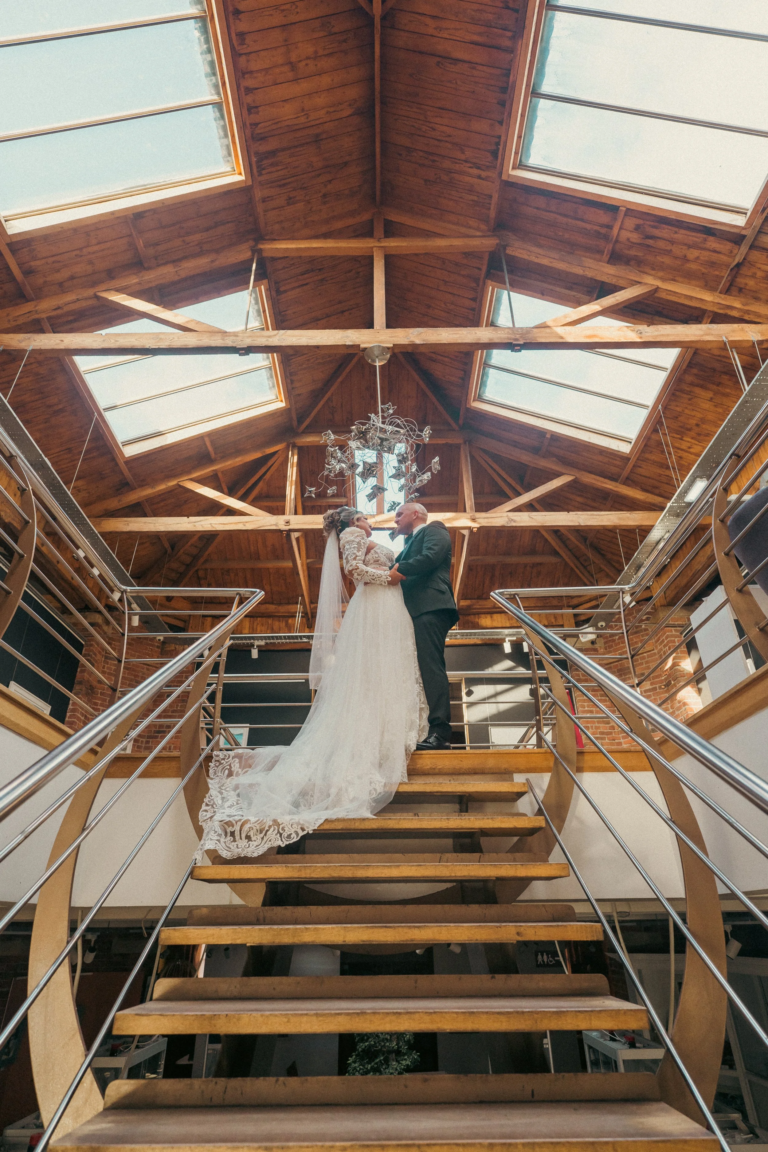 A bride and groom holding hands and gazing at each other on a staircase in a rustic, wooden-ceiling venue with skylights and a chandelier.