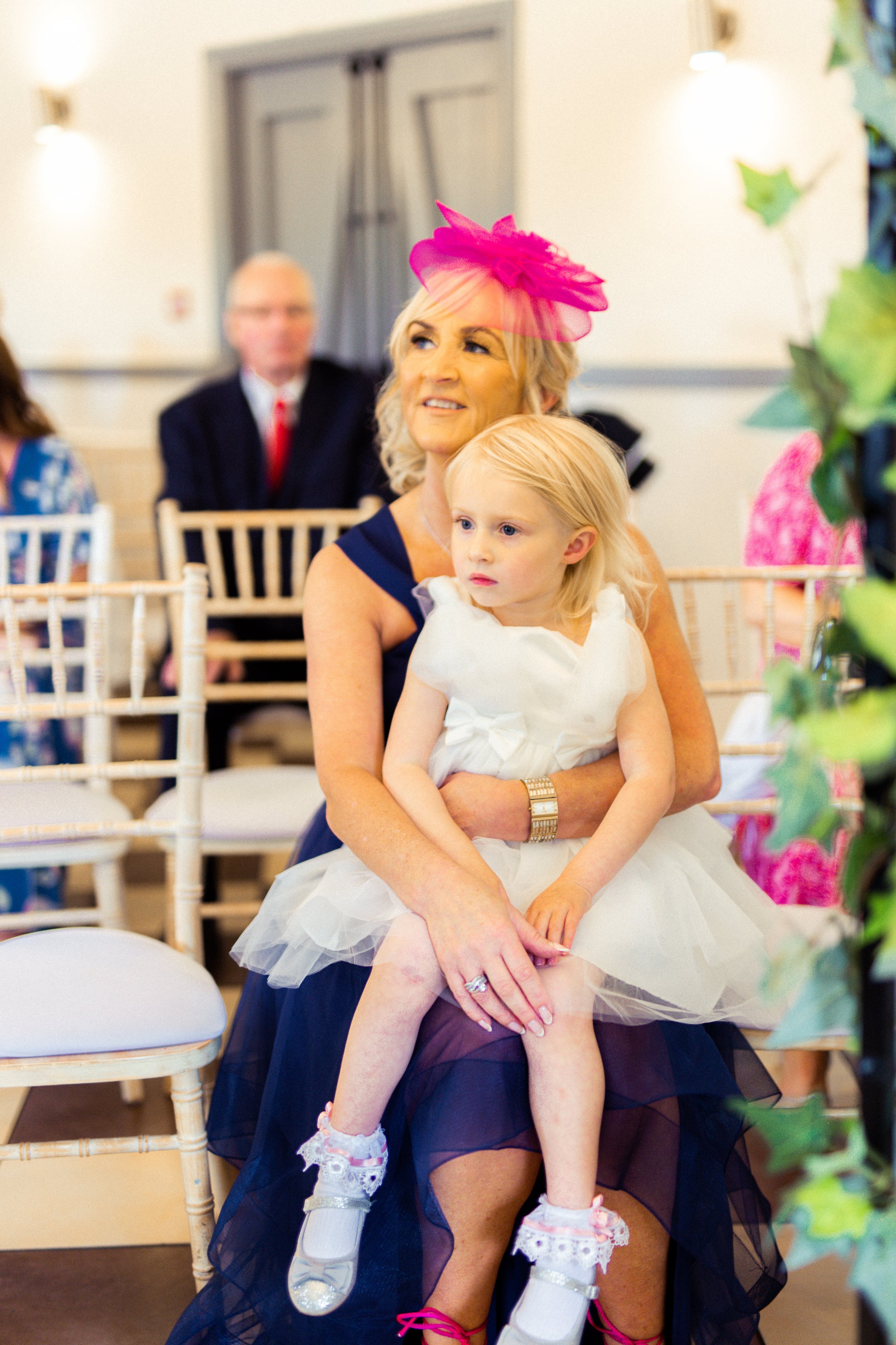 A woman in a navy dress and a pink fascinator hat sits on a chair, holding a young girl with blonde hair in a white dress on her lap at a social gathering or wedding.