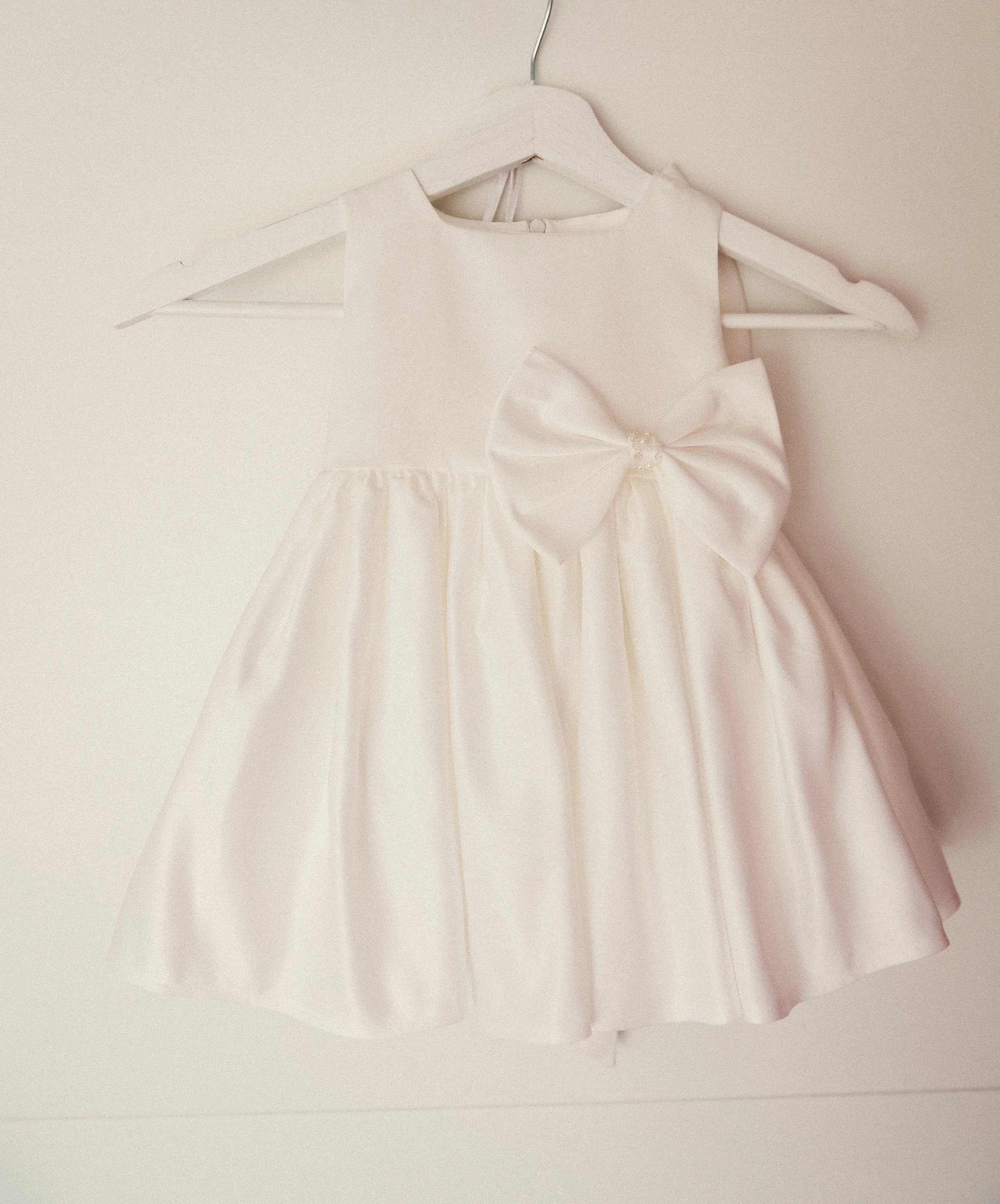 A white satin dress with a large bow on the front, hanging on a white hanger against a plain background.