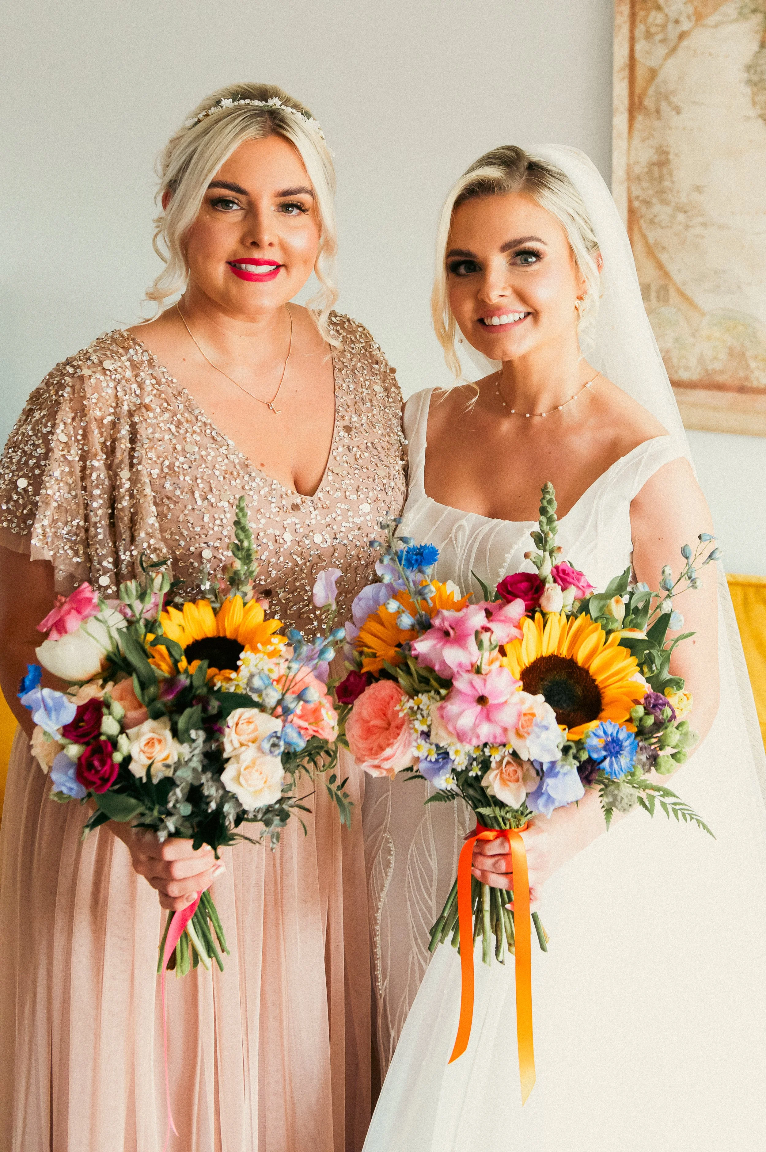 Two women, one in a glittery dress and the other in a wedding dress, holding colorful bouquets of flowers, smiling indoors.