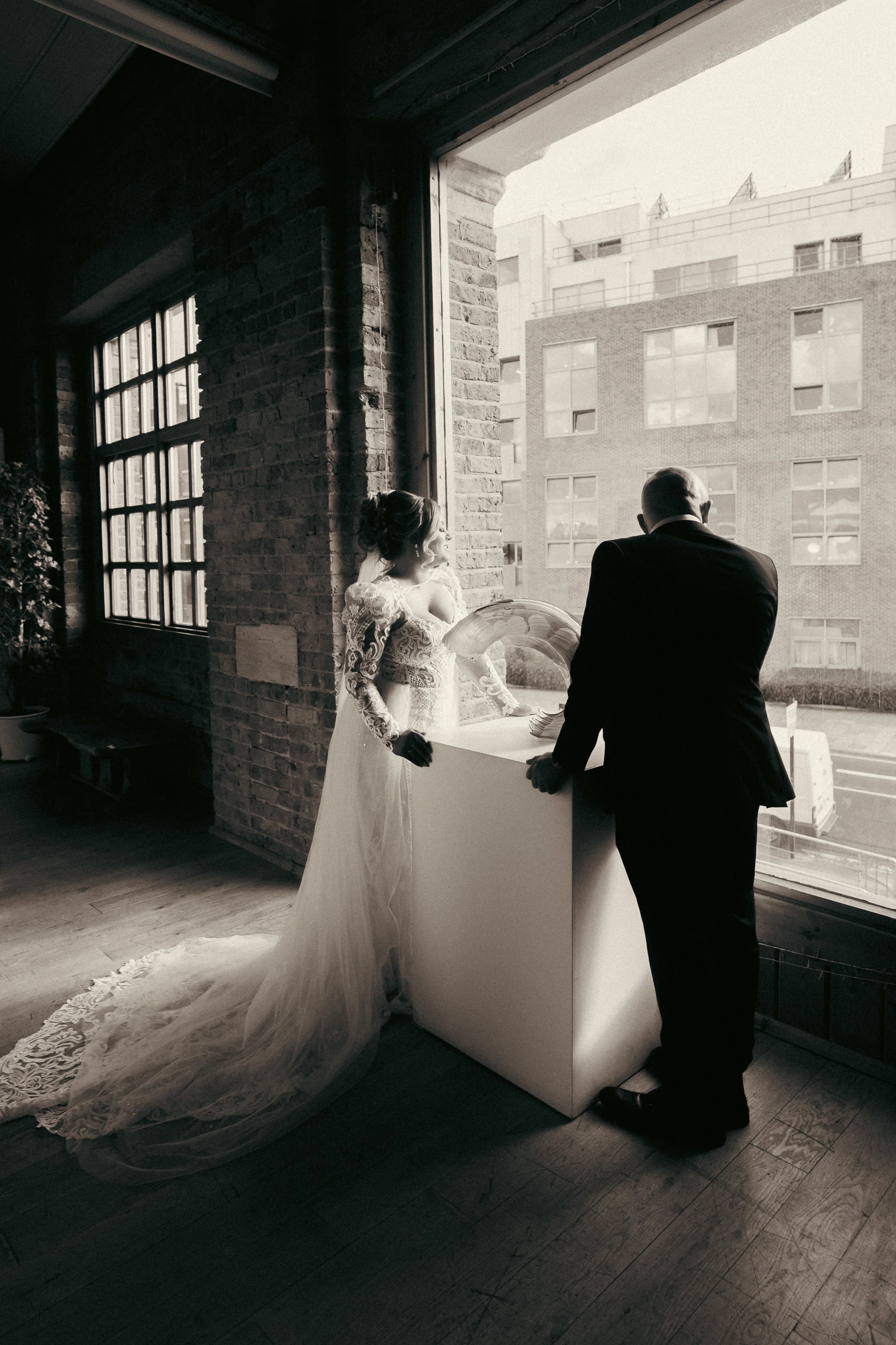 Couple dressed in wedding attire standing near a large window, with the woman in a lace wedding gown and veil, and the man in a dark suit, looking outside.