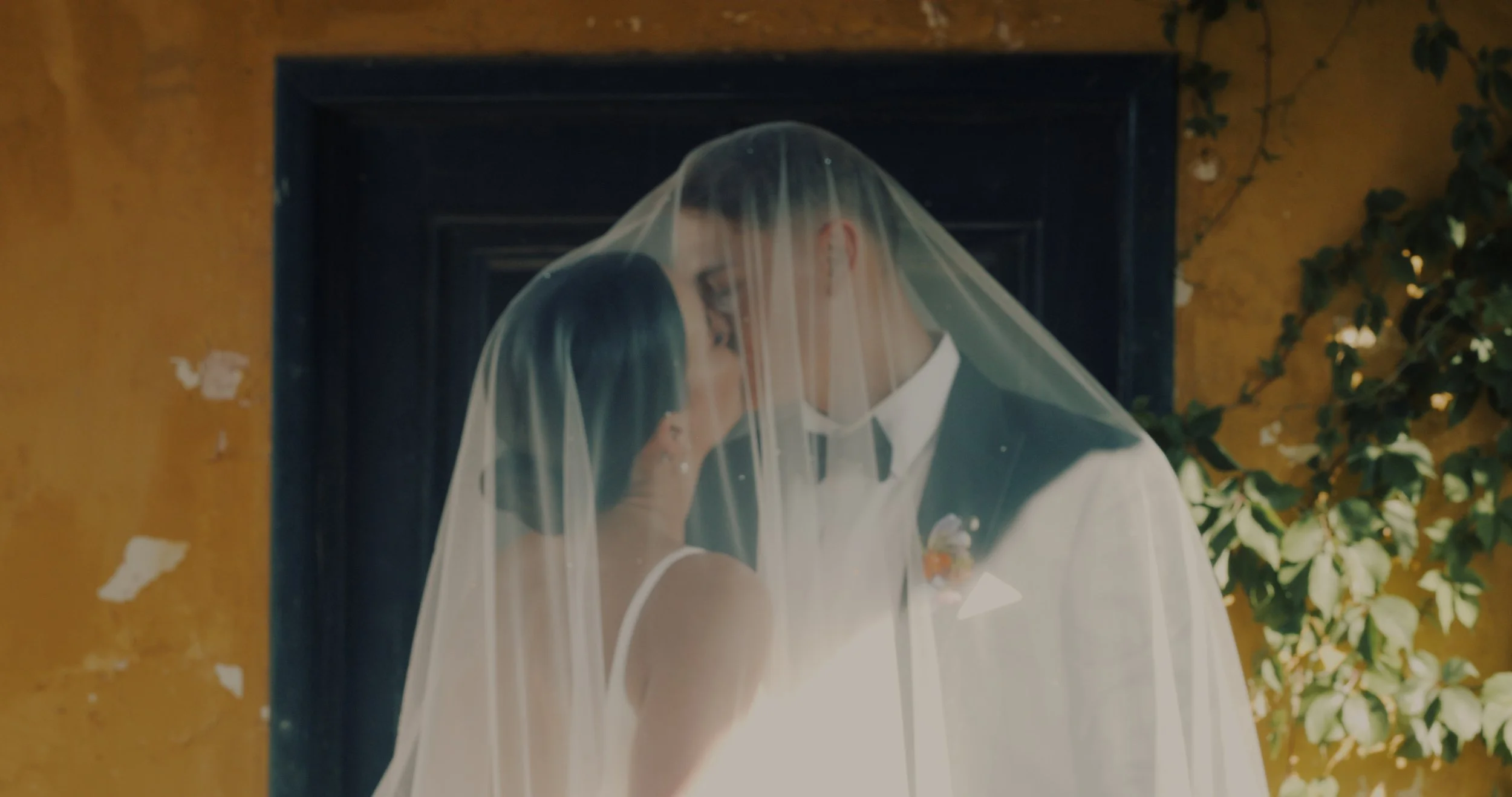 A bride and groom with their foreheads touching, sharing a tender moment under a veil inside a rustic building with a yellow wall and greenery.
