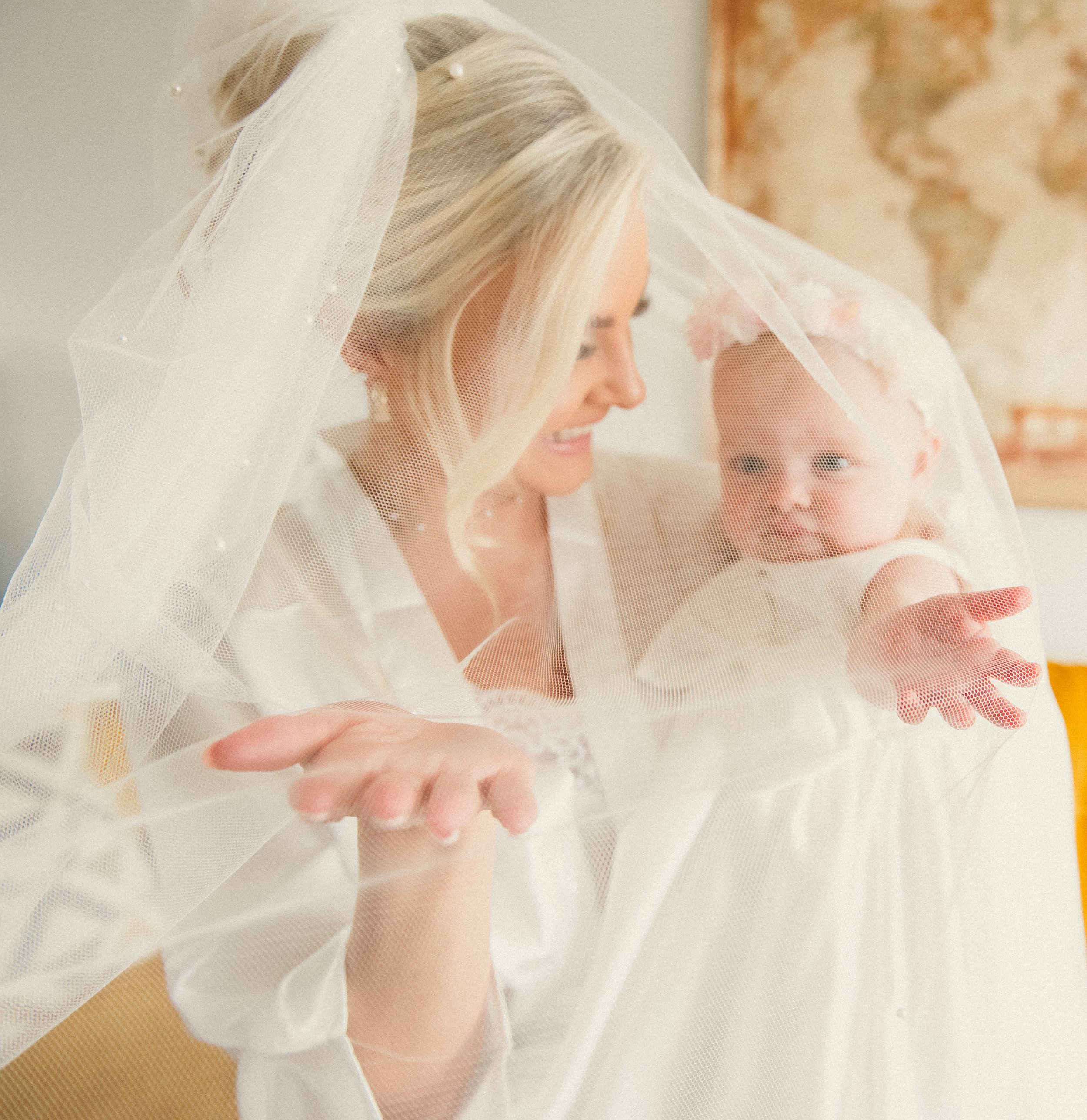A smiling woman in a wedding dress and veil interacts with a baby girl who is also dressed in white and has a floral headband, as they sit on a yellow couch indoors.