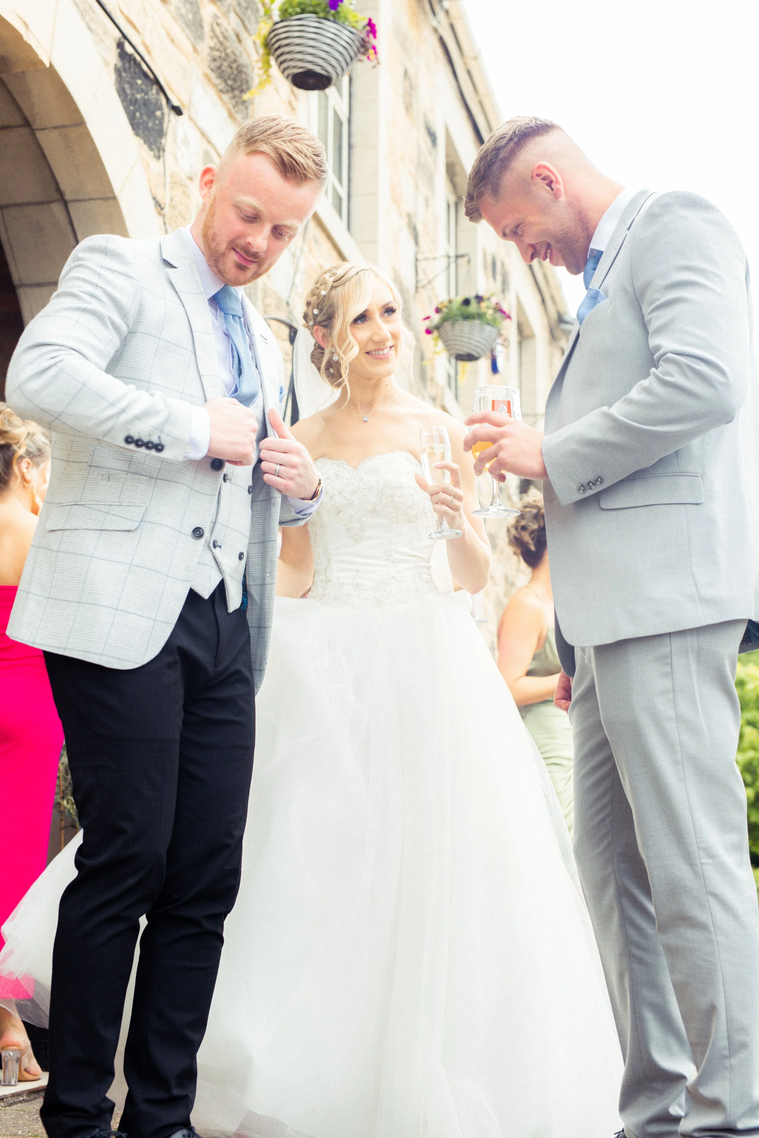 A bride and two grooms are outdoors, celebrating with glasses of champagne during a wedding event. The bride, in a white wedding gown, stands between the two men dressed in suits. The background shows a stone building with hanging flower baskets.